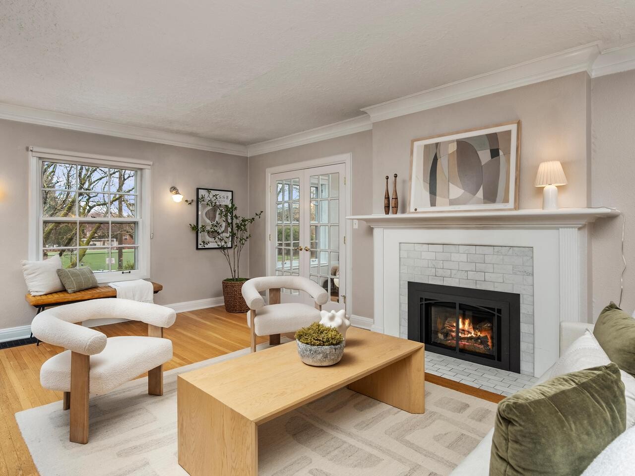 A modern living room with light walls, wood floors, abstract art, a lit fireplace, two white chairs, a light wood coffee table, a white sofa with green pillows, and large windows showing trees outside.