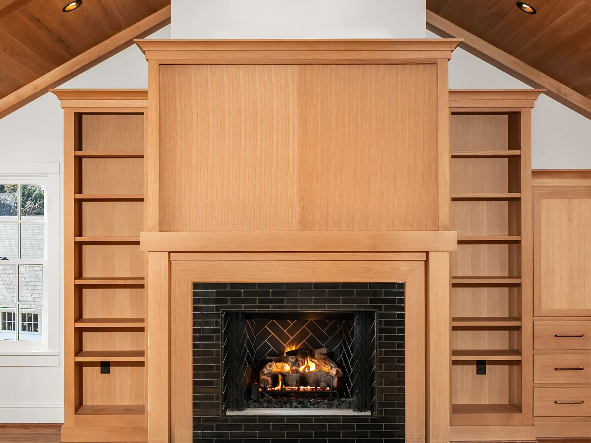A modern living room with wood-paneled vaulted ceiling, built-in shelves and cabinets, and a central fireplace with a black tile surround. Warm sunlight enters through a window on the left.