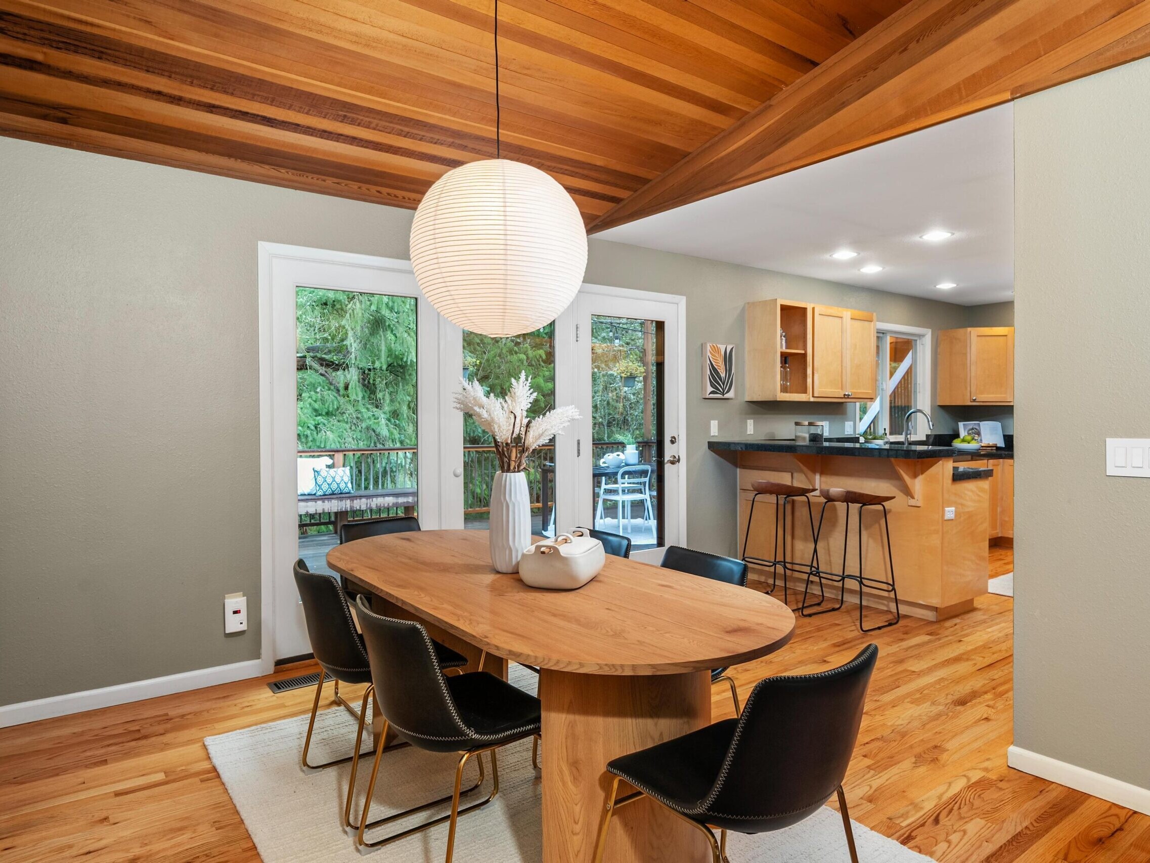 A modern dining area with a wooden oval table, black chairs, a round pendant light, and a vase with flowers. Behind, there’s a kitchen with wooden cabinets and a bar, and glass doors open to a green outdoor view.