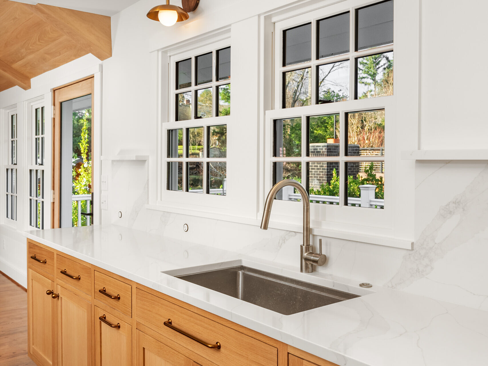 A modern kitchen with wooden cabinets, a white marble countertop and backsplash, a stainless steel sink, large windows, and two brass wall sconces above the sink area.