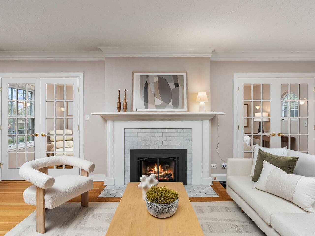 A modern living room with a lit fireplace, neutral-colored furniture, a wooden coffee table with decorative objects, and glass double doors on either side of the fireplace. Artwork and vases decorate the mantel.