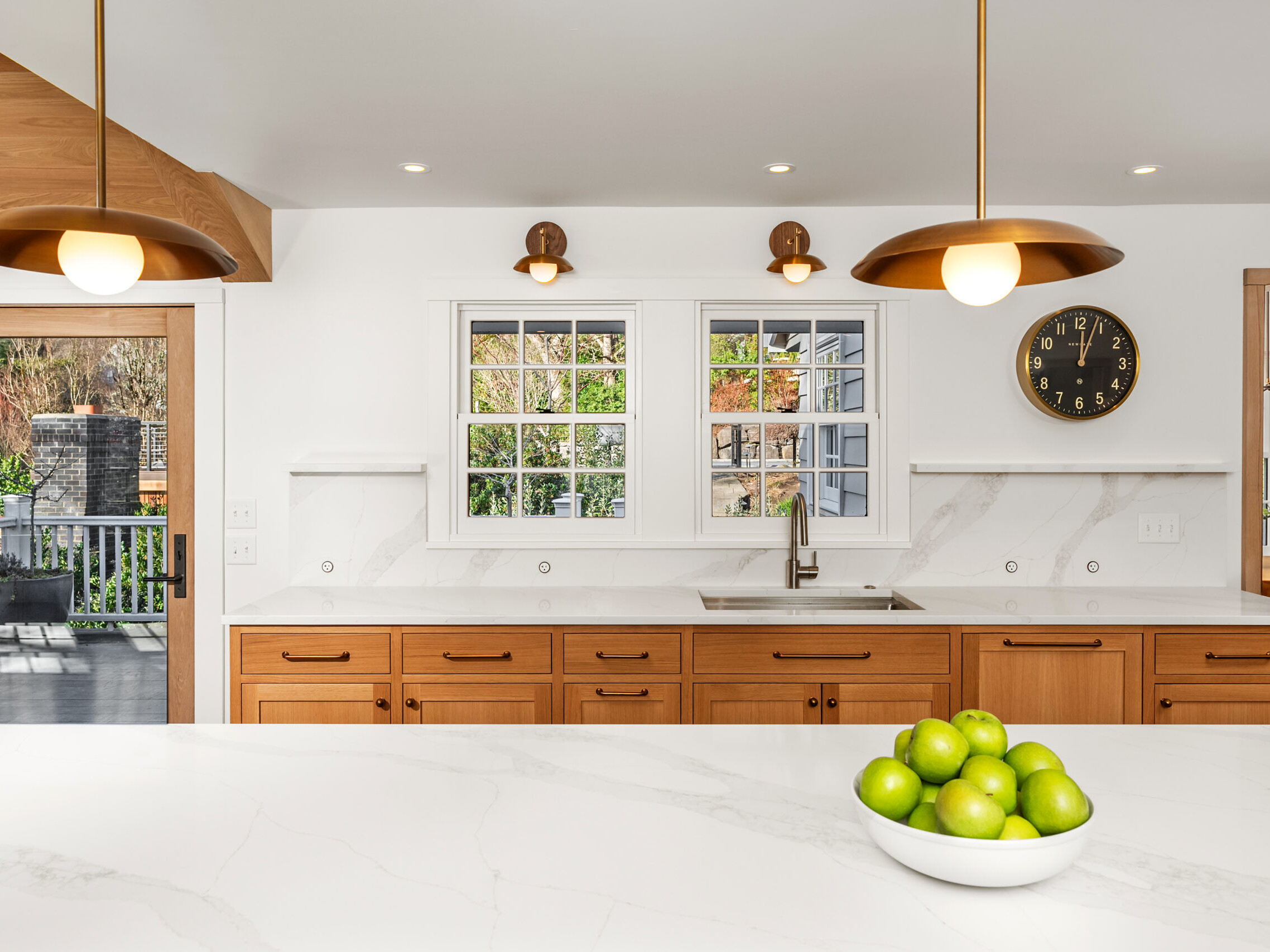 Modern kitchen with wooden cabinets, white marble countertops, gold pendant lights, a sink under a window, and a bowl of green apples on the island; a black wall clock and outdoor view are visible.