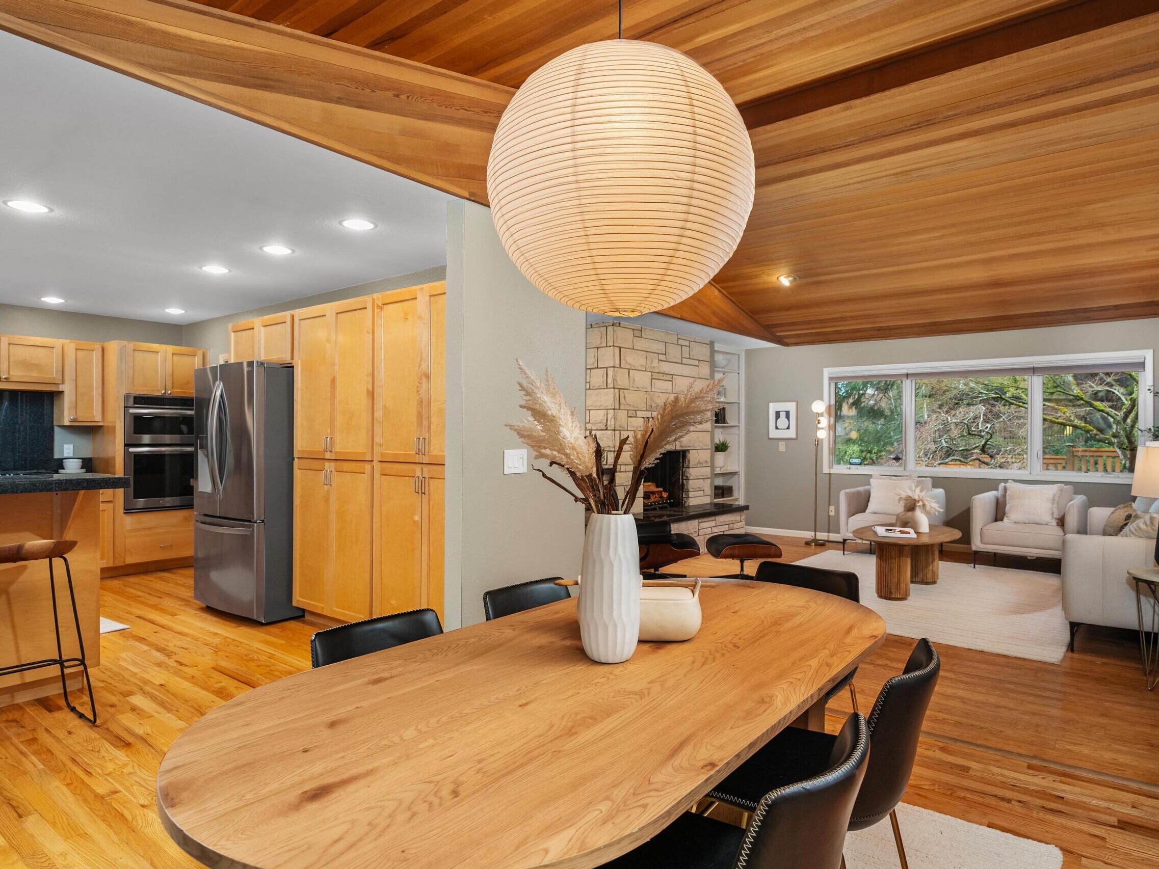 A modern open-concept kitchen, dining, and living area with wood floors and ceilings, light wood cabinets, a round dining table, neutral decor, and large windows letting in natural light.