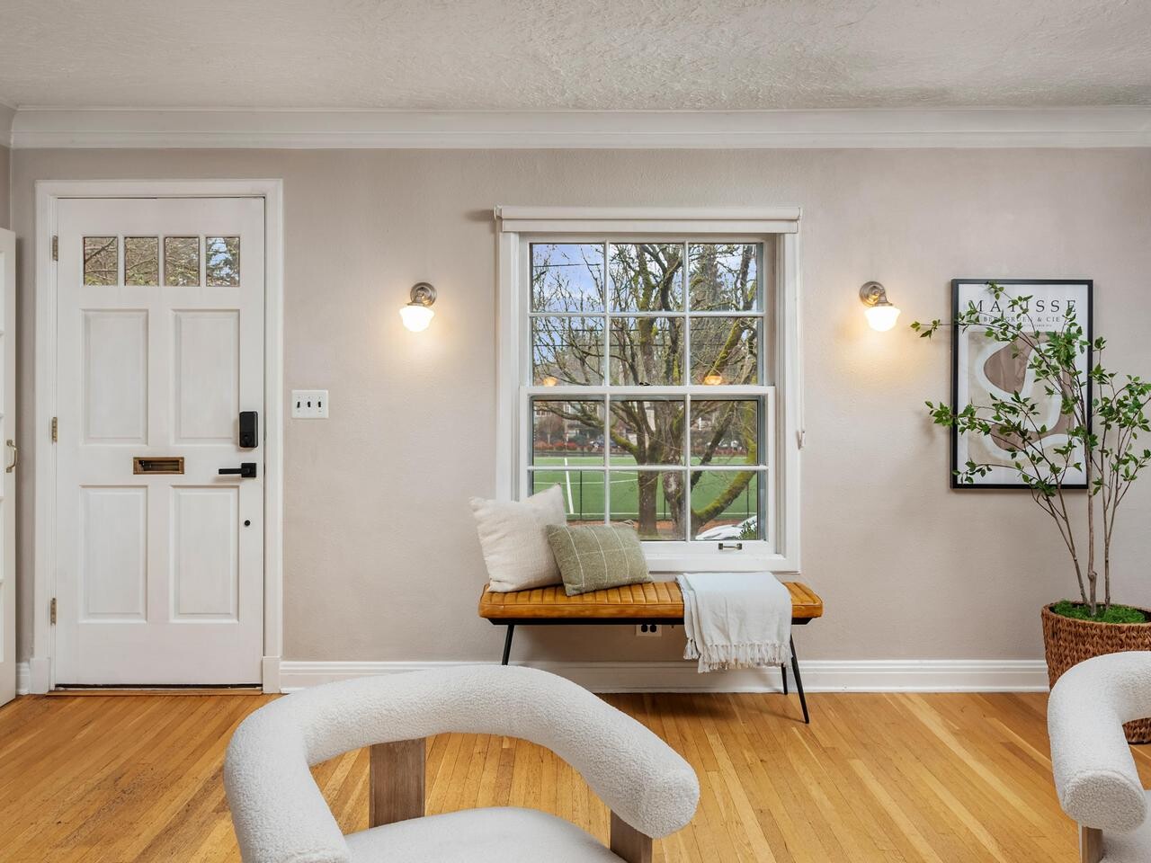 Bright living room with light wood floors, a white front door, and a window showing a view of trees and grass. A bench with cushions sits under the window. A potted plant and framed art are on the right wall.