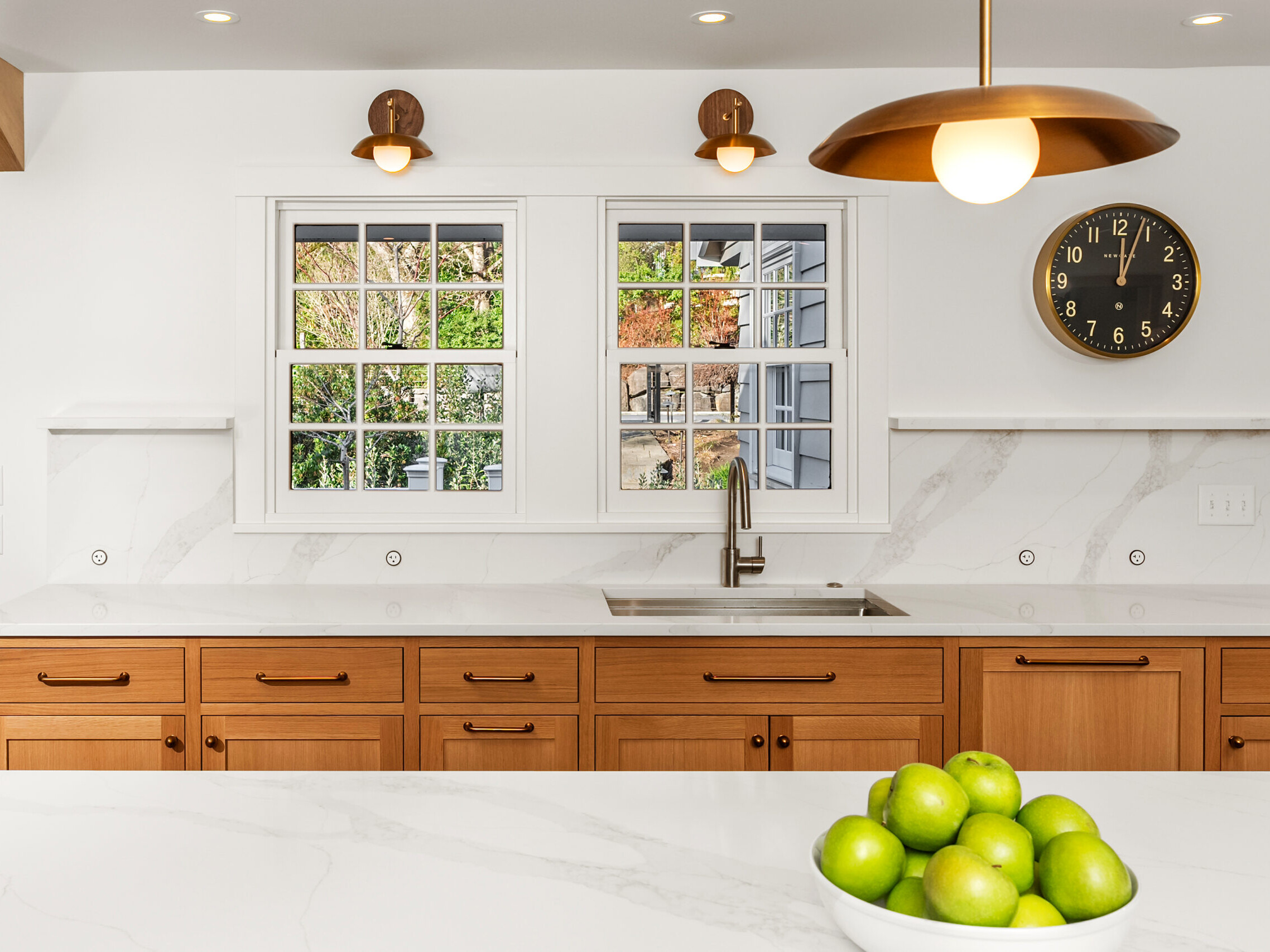 Modern kitchen with white marble countertops, light wood cabinets, a sink beneath double windows, brass light fixtures, a black wall clock, and a bowl of green apples on the counter.