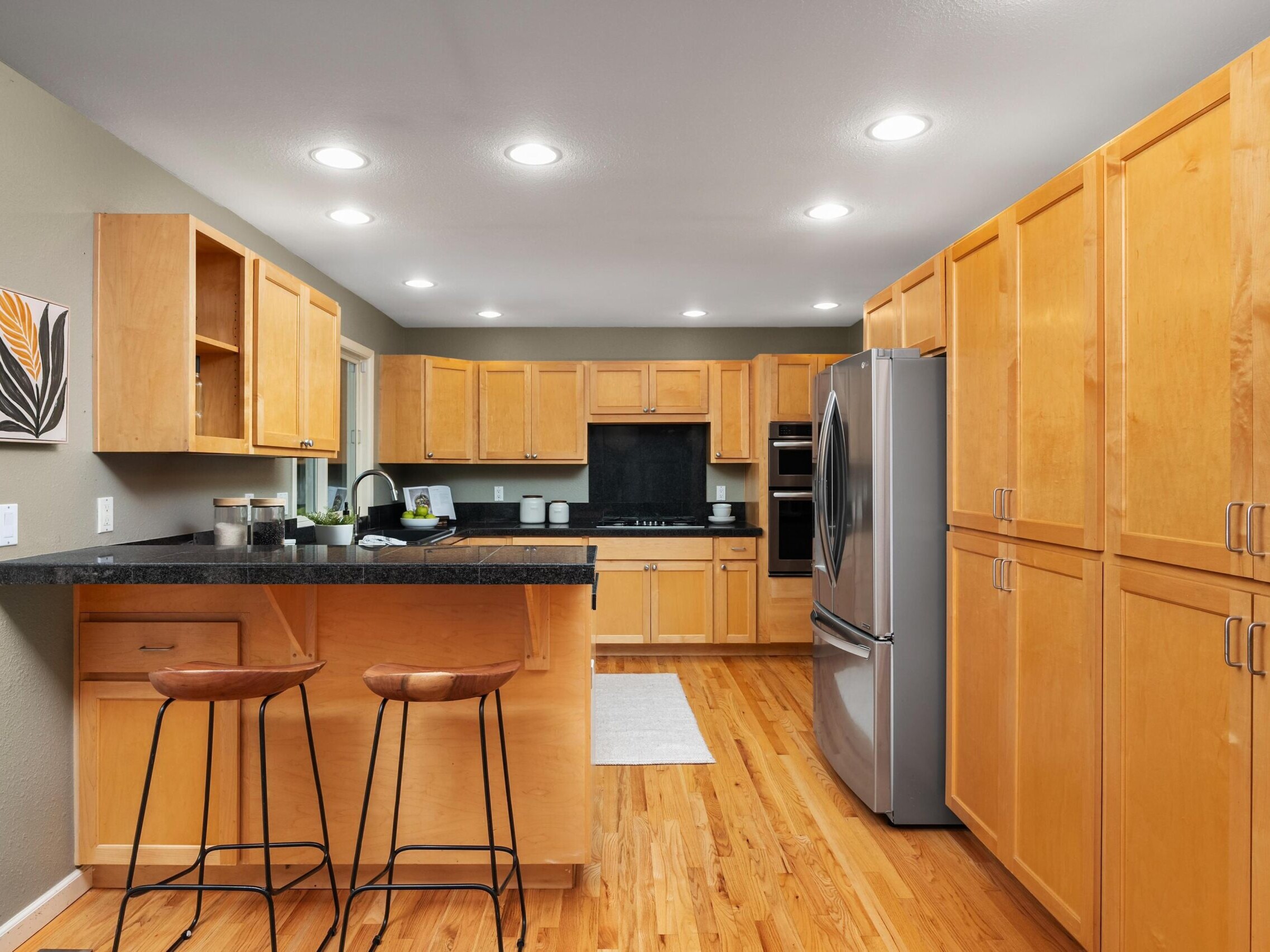 Modern kitchen with light wood cabinets, black countertops, stainless steel appliances, two brown barstools at an island, and a wall art piece with a leaf design; wood flooring and recessed ceiling lights.