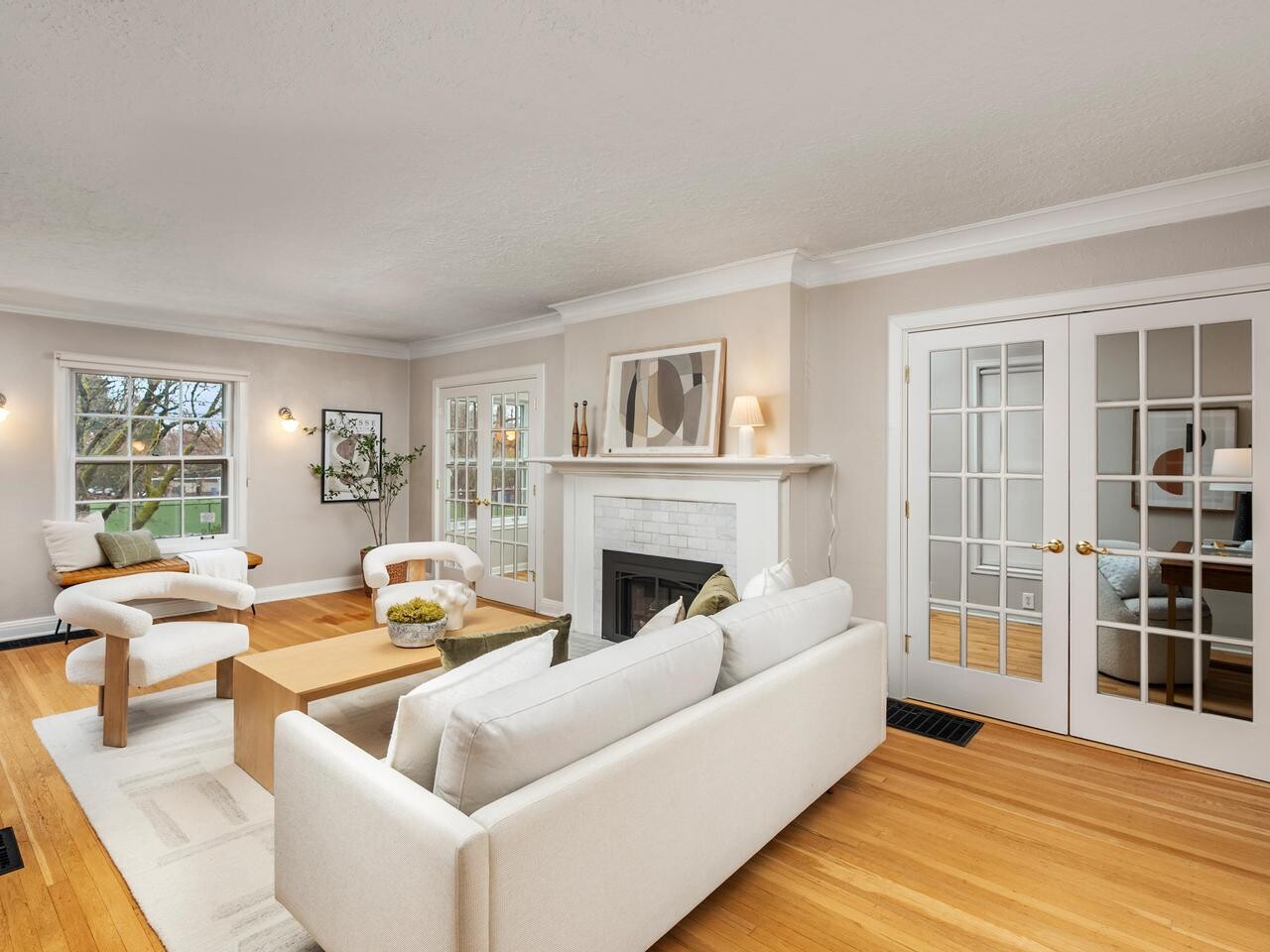 Bright living room with light wood floors, white walls, a white sofa and chairs around a wooden coffee table, a fireplace with art above it, large windows, and glass-paneled French doors.
