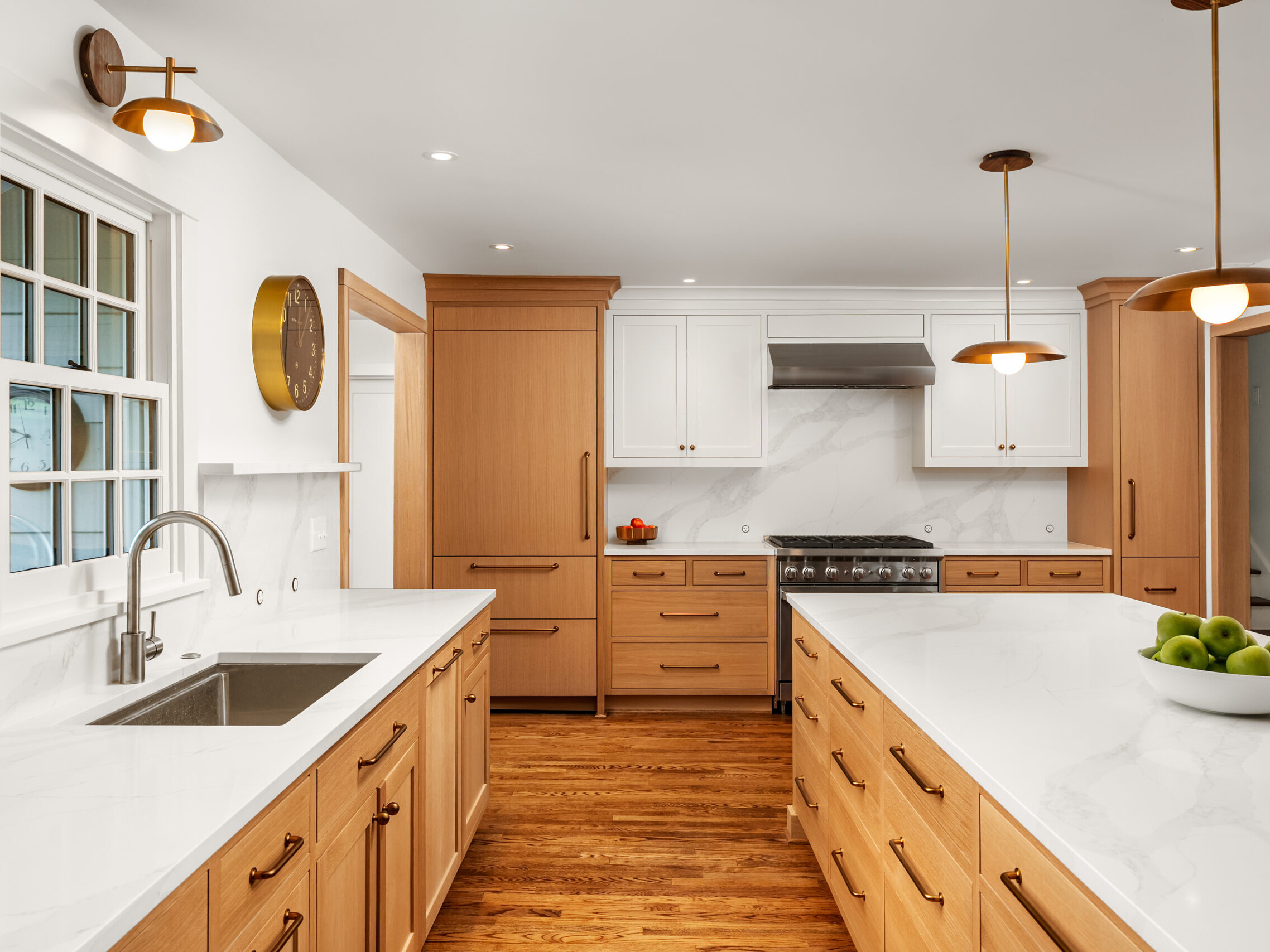 A modern kitchen with light wood cabinets, white marble countertops, stainless steel appliances, and pendant lights. A bowl of green apples sits on the island; a window and a clock are on the left wall.