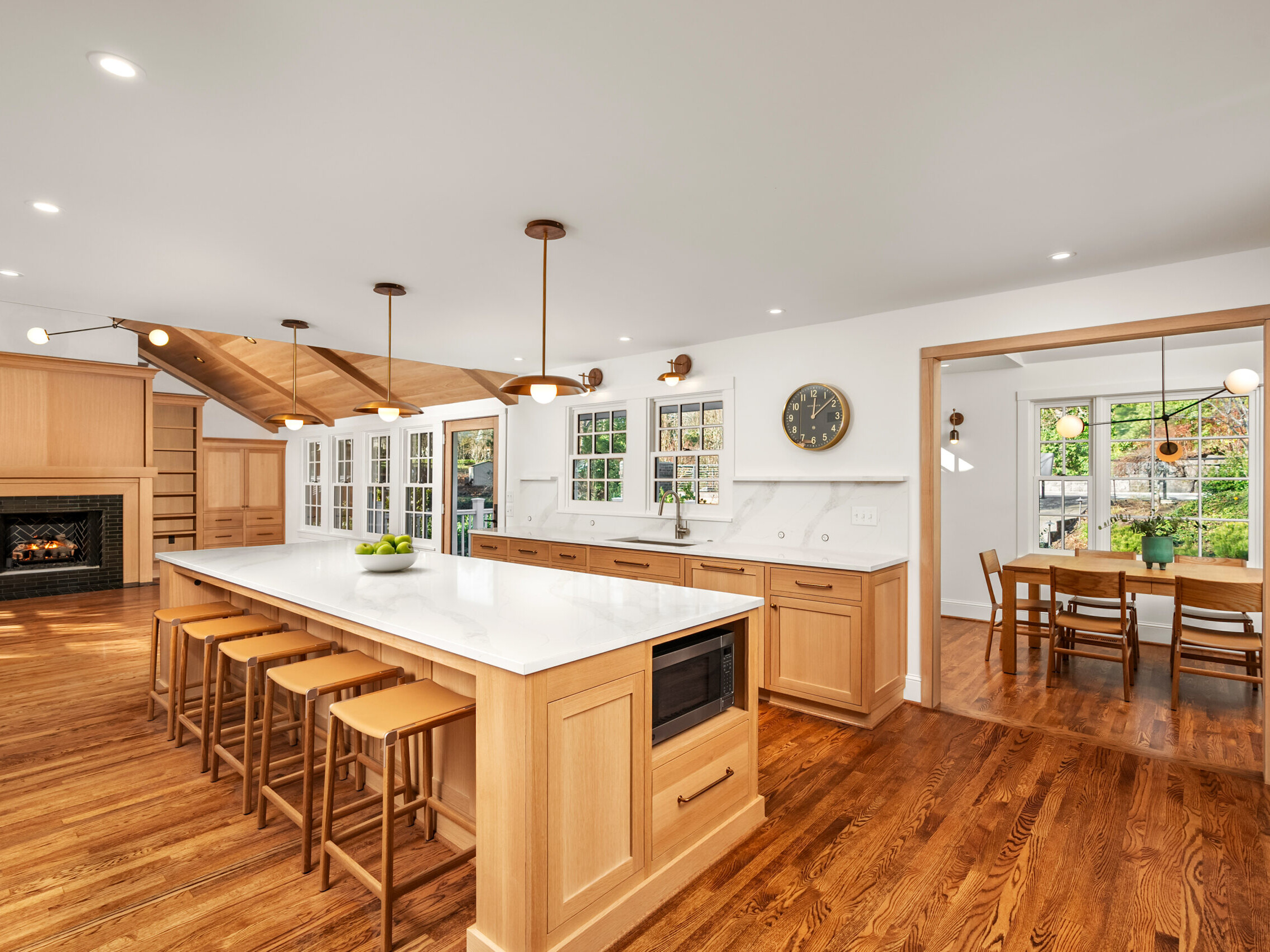 Spacious, modern kitchen with light wood cabinets, white countertops, a large island with stools, and pendant lights; adjacent to a dining area with a wooden table and large windows.