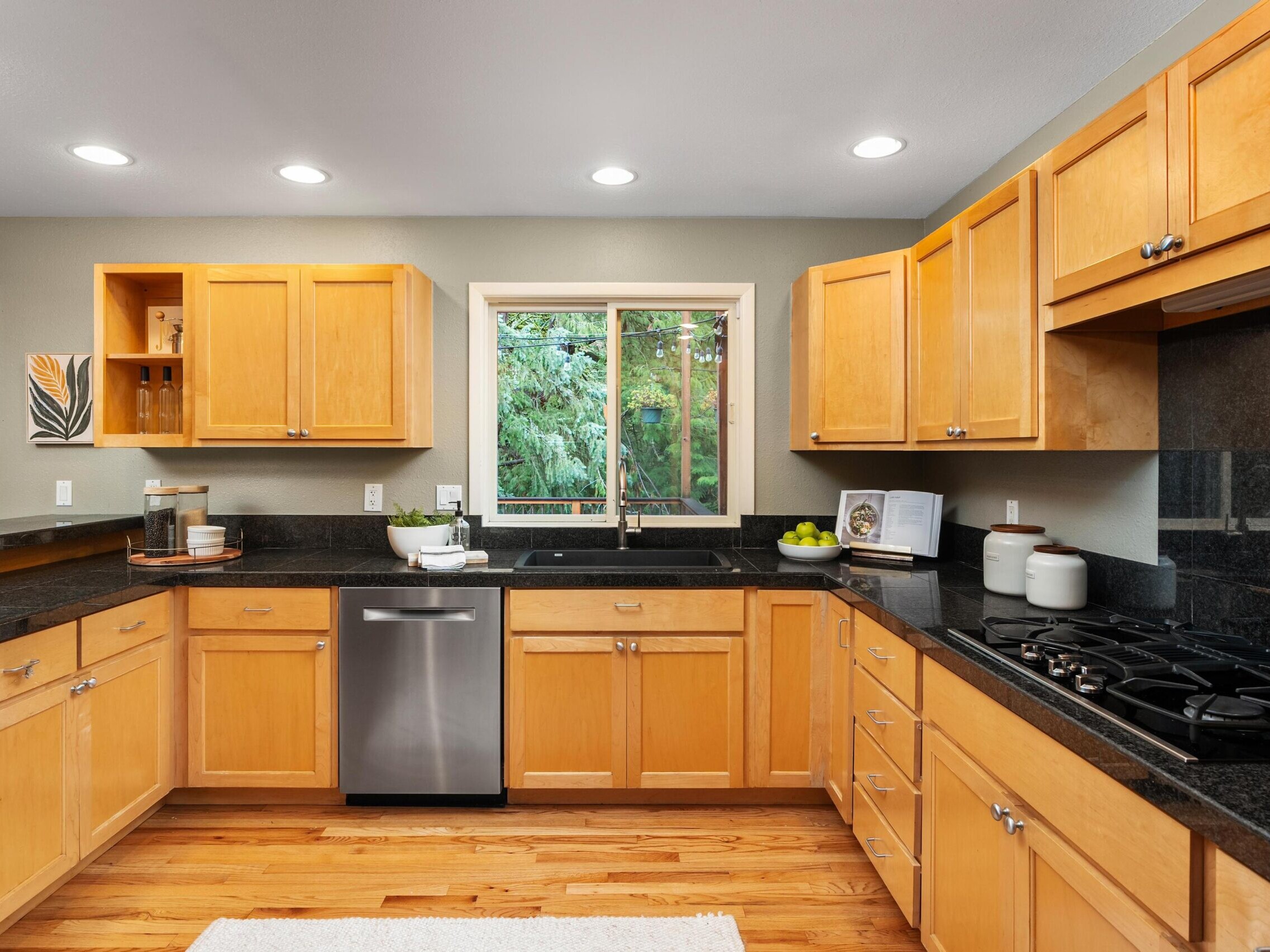 Modern kitchen with light wood cabinets, black countertops, stainless steel dishwasher, gas stove, and a window overlooking green trees. Decorative items and kitchen essentials are neatly arranged on the counters.