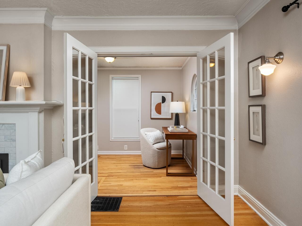 A bright living room with light wood floors, white French doors, a white sofa, and a desk with a chair in the adjacent room. Modern art, a lamp, and framed pictures decorate the neutral-colored space.