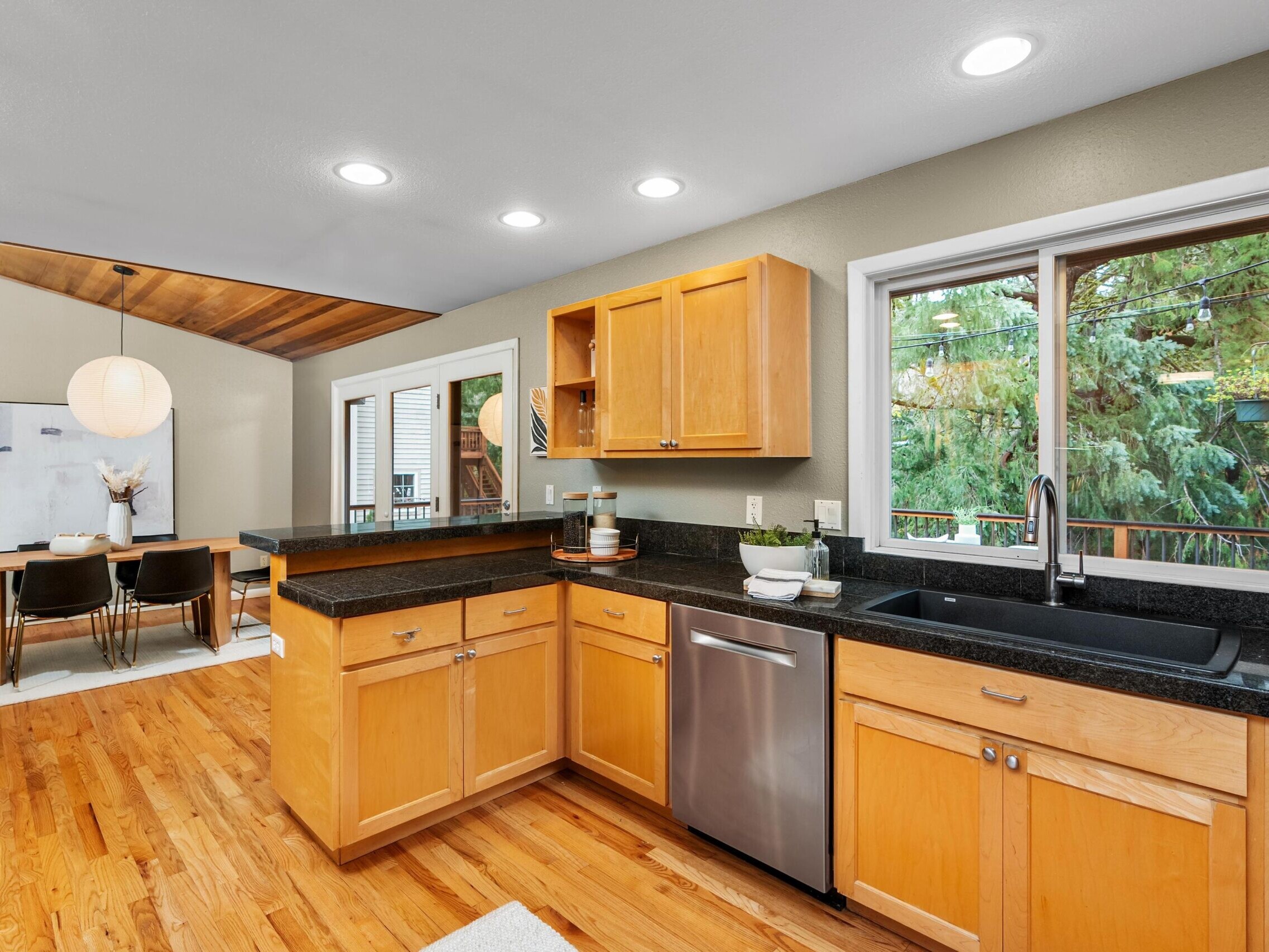 Modern kitchen with light wood cabinets, black countertops, stainless steel appliances, and a window above the sink. The adjacent dining area features a wooden table, modern chairs, and a large round hanging light.