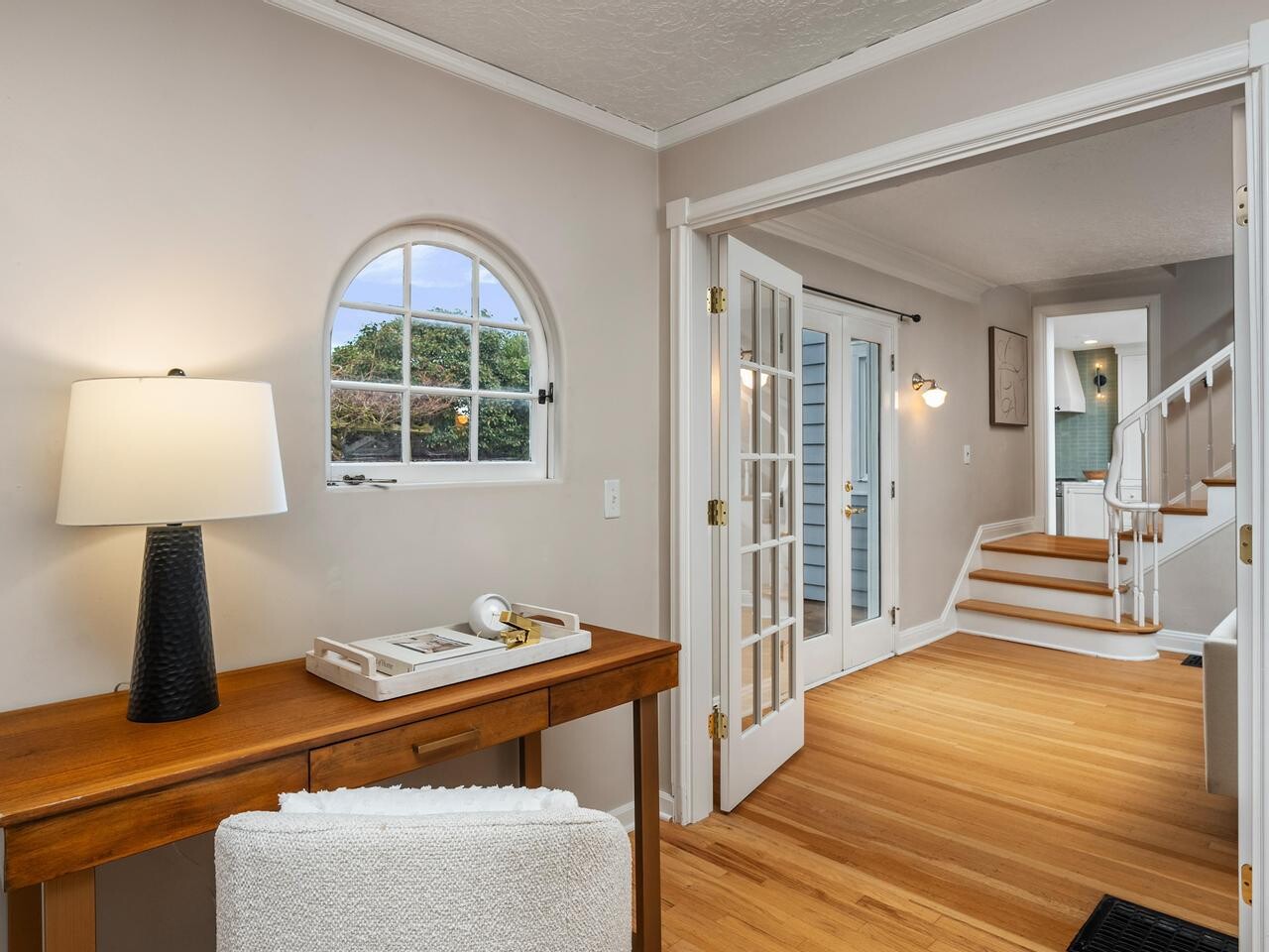 A cozy home entryway with a wooden desk and chair, a lamp, a small arched window, and an adjoining room with French doors and a staircase leading upstairs. The space features light walls and hardwood floors.