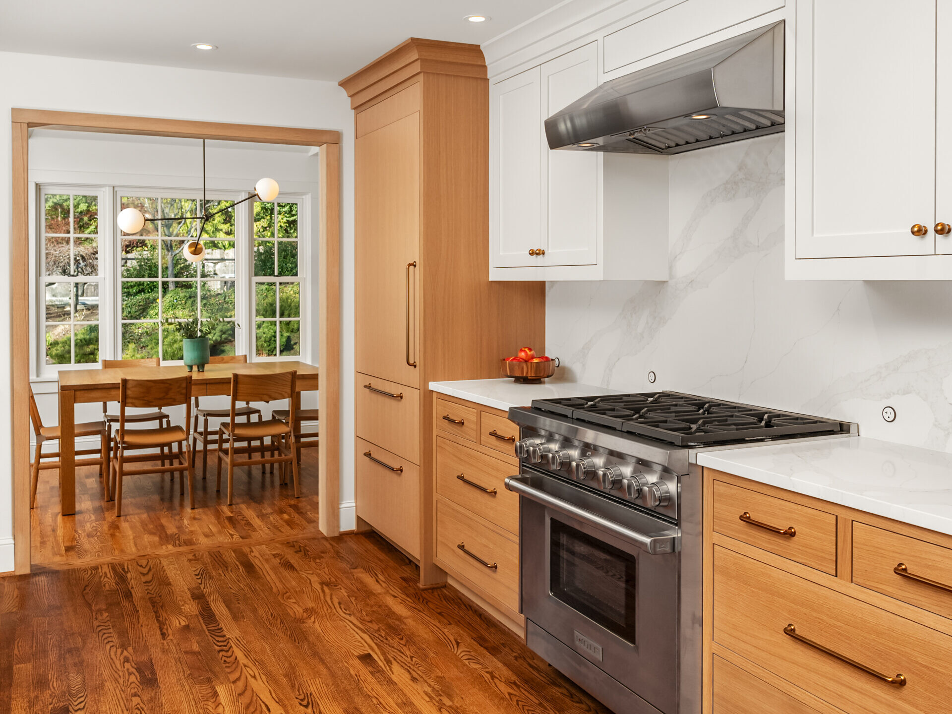 A modern kitchen with white upper cabinets, light wood lower cabinets, stainless steel stove, and hood. The kitchen opens to a dining area with a wooden table, six chairs, and large windows overlooking greenery.