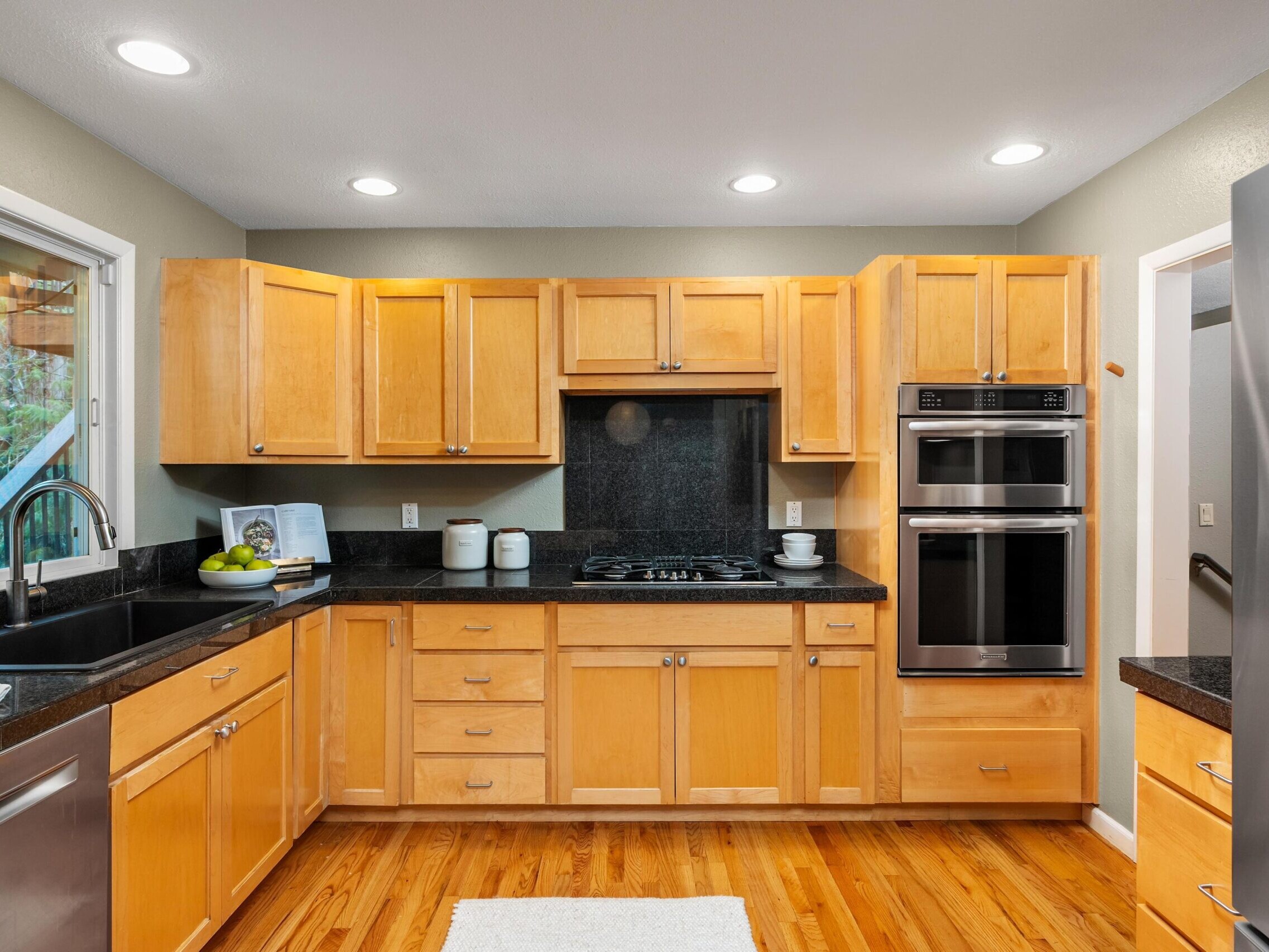 Modern kitchen with light wood cabinets, black countertops, stainless steel appliances, a double oven, gas cooktop, and a large sink under a window. The floor is wood and recessed ceiling lights are on.