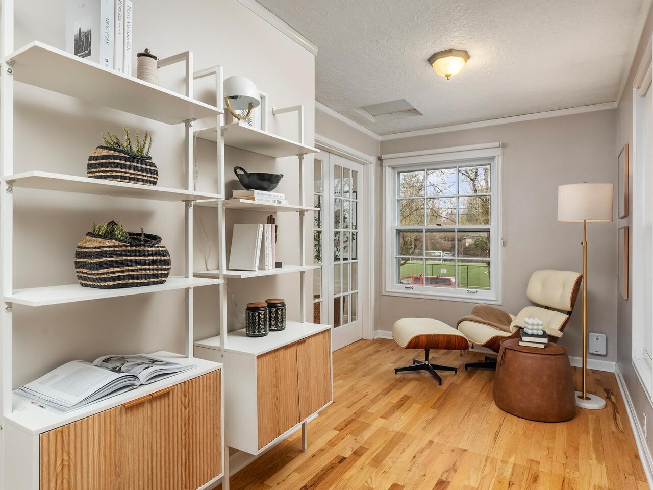 A cozy reading nook with a white lounge chair and ottoman, next to a tall white bookshelf holding books, decor, and plants. Large windows and glass doors let in natural light, and a floor lamp stands beside the chair on a wooden floor.