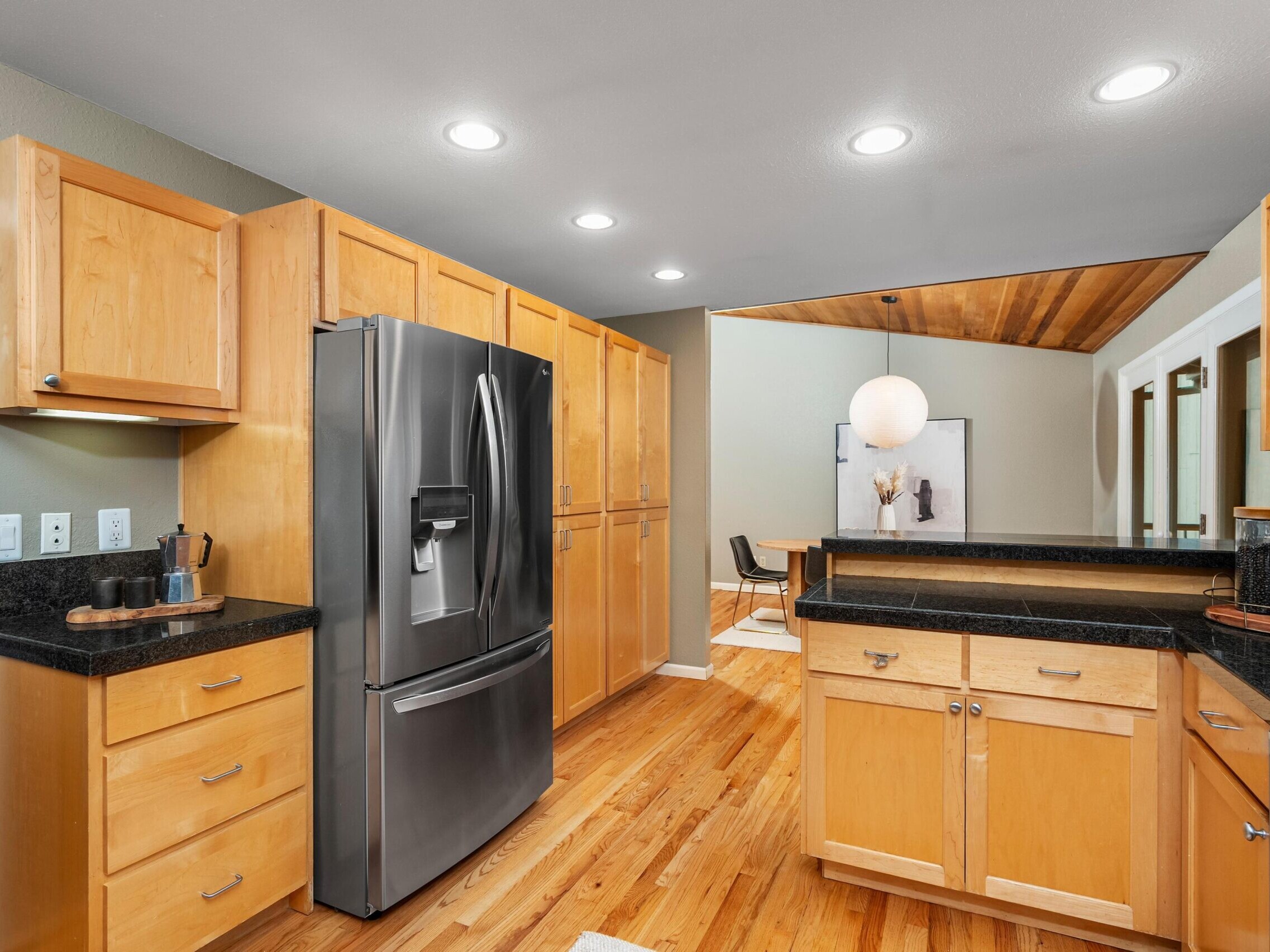 Modern kitchen with light wood cabinets, black countertops, stainless steel refrigerator, and wood flooring. There is a small dining area visible in the background with a round white pendant light.