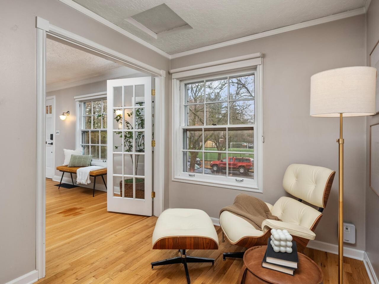 A cozy reading nook with an Eames lounge chair and ottoman beside a window, a round side table with decor, a modern floor lamp, wood floors, and an adjacent room with French doors.