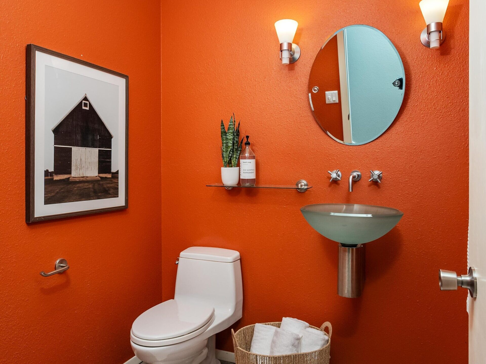 A modern bathroom with bright orange walls, a white toilet, a round mirror above a glass sink, two wall lights, a framed barn photo, a small plant on a glass shelf, and a basket with towels on a stone floor.
