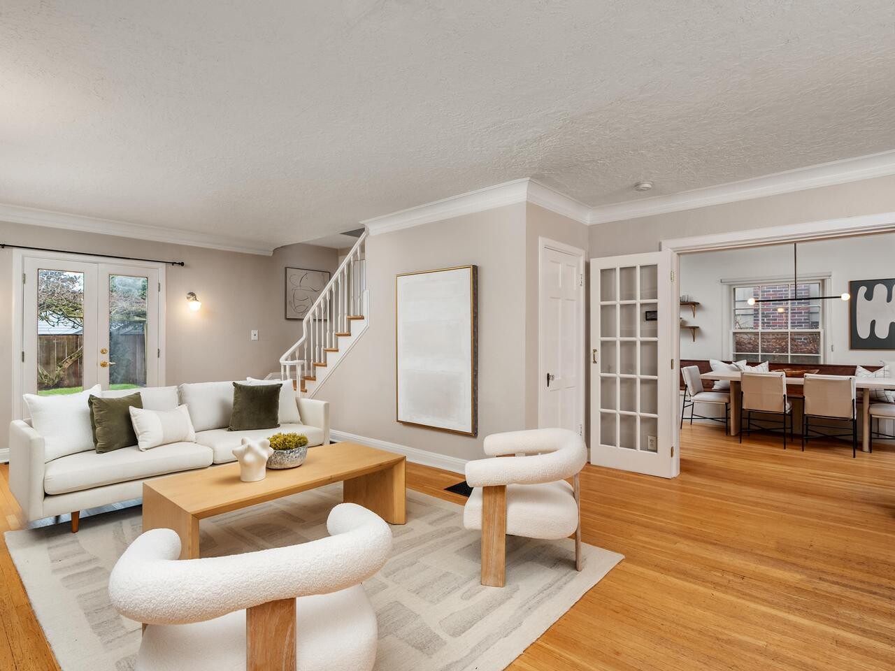 A modern living room with neutral tones, featuring a white sofa, two white chairs, a wooden coffee table, hardwood floors, and an open doorway leading to a dining area with a large window.