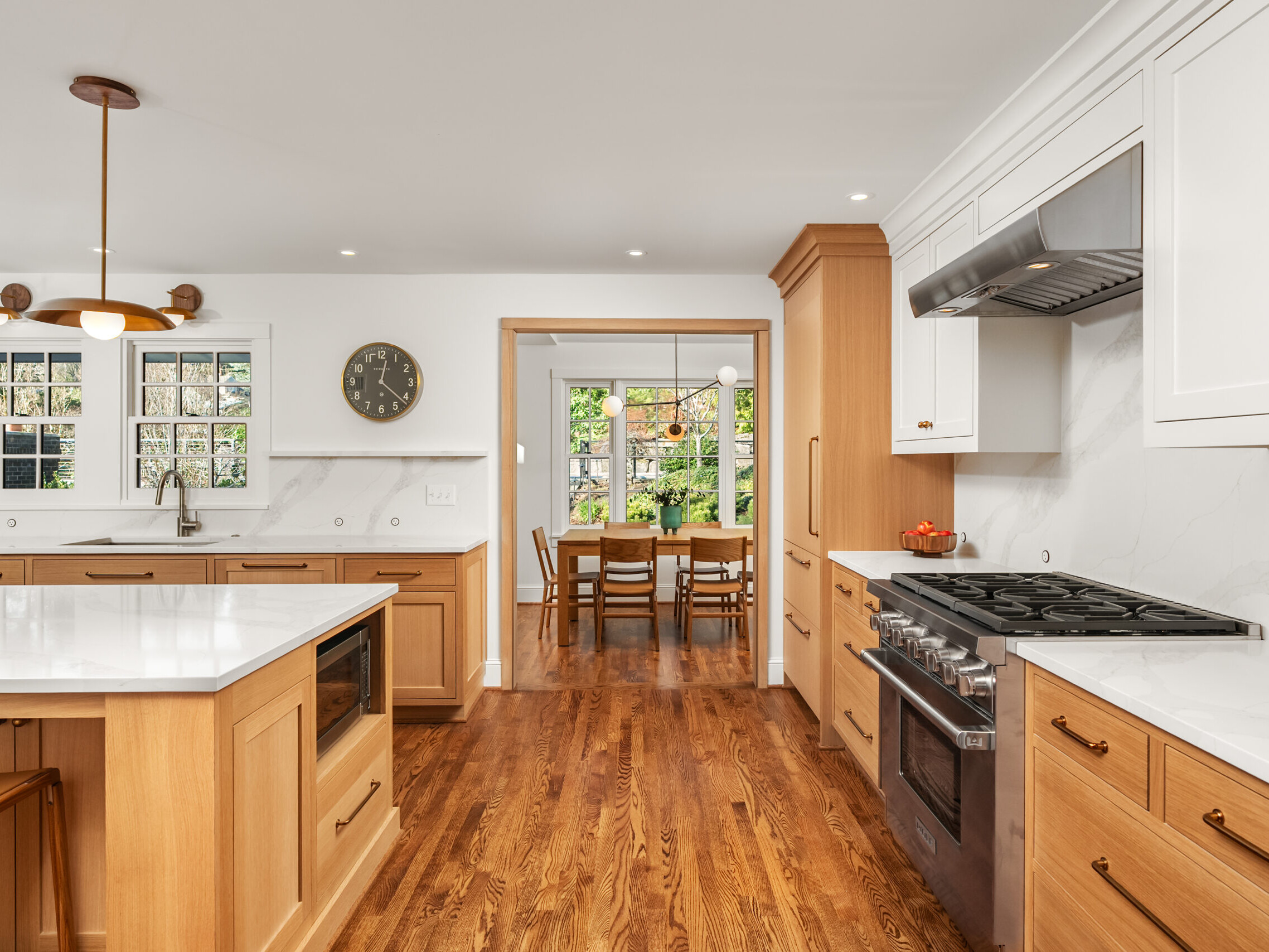 A spacious, modern kitchen with wood cabinets, white countertops, stainless steel appliances, and an island. Natural light fills the room, and a dining area is visible through an open doorway in the background.
