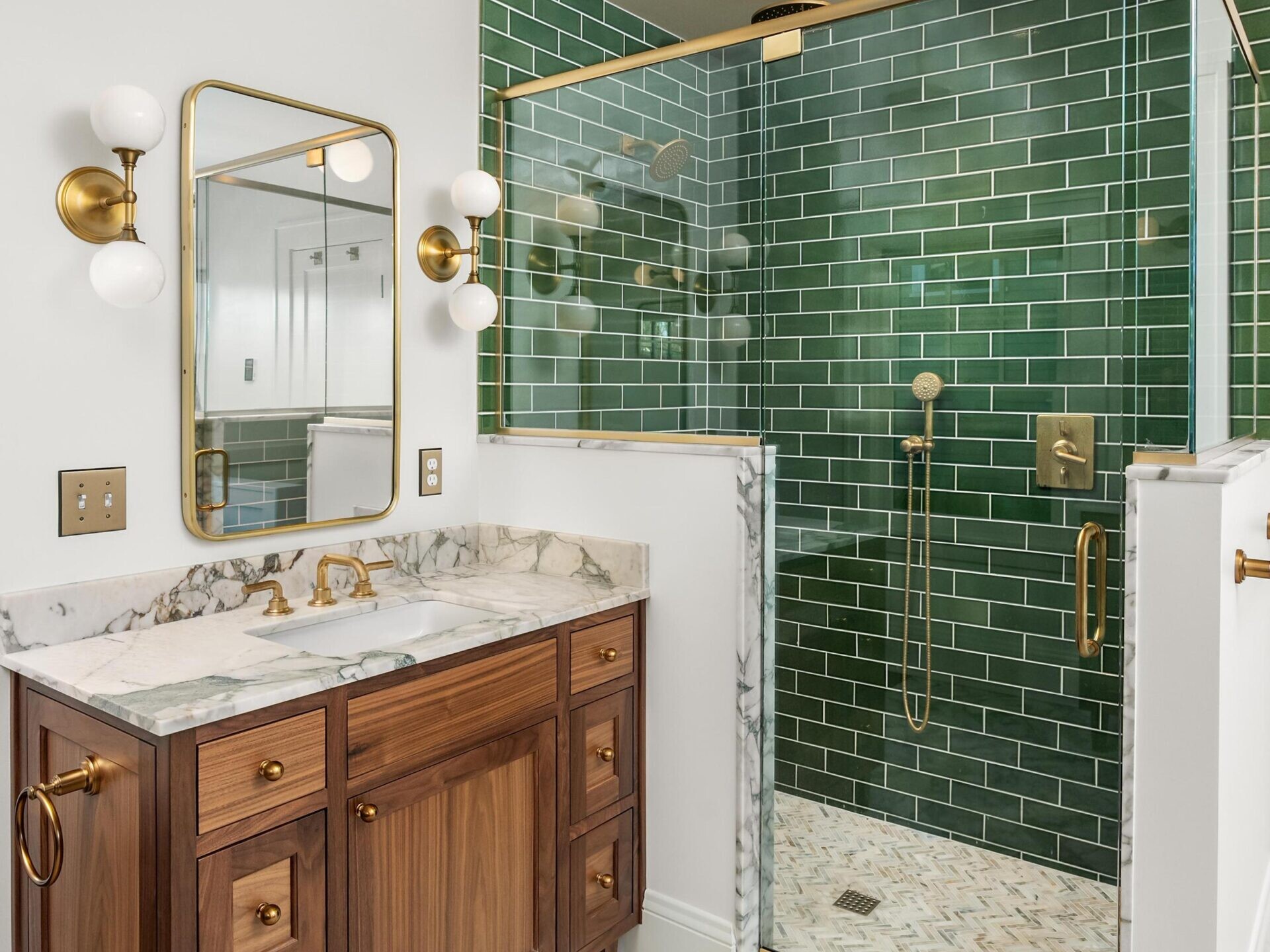 Modern bathroom with a marble floor, wooden vanity with marble countertop, gold fixtures, a rectangular mirror, and a glass-enclosed shower featuring green subway tiles and gold accents.