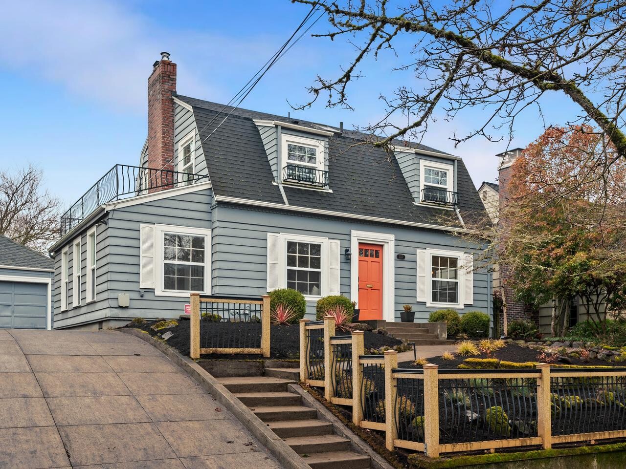 A two-story gray house with white trim, a steep sloped driveway, an orange front door, and a small fenced front yard with shrubs and black mulch. A detached garage is visible on the left.