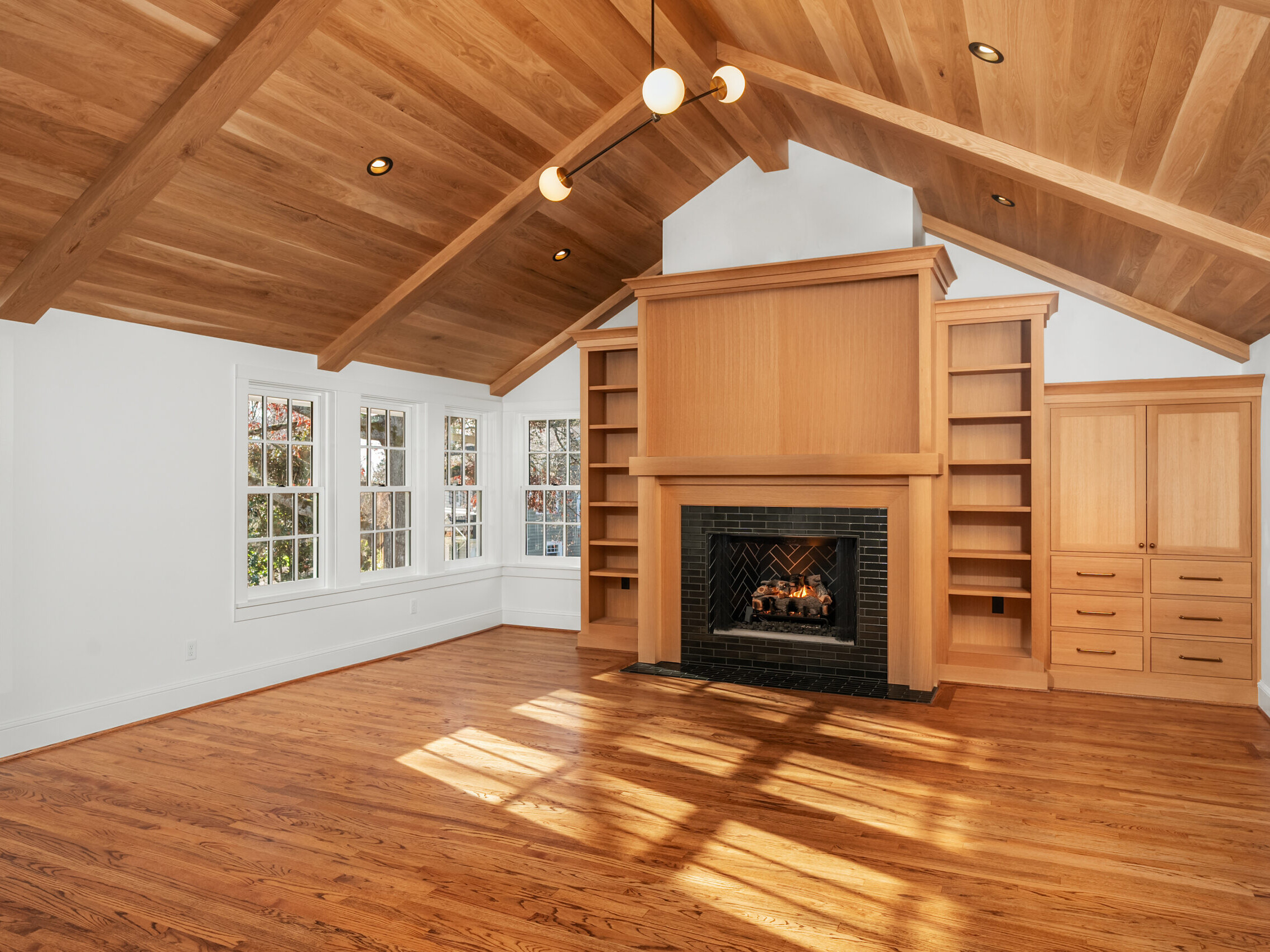A spacious living room with a vaulted wood ceiling, hardwood floors, built-in shelves and cabinets, a large fireplace with a black surround, and several windows letting in natural light.