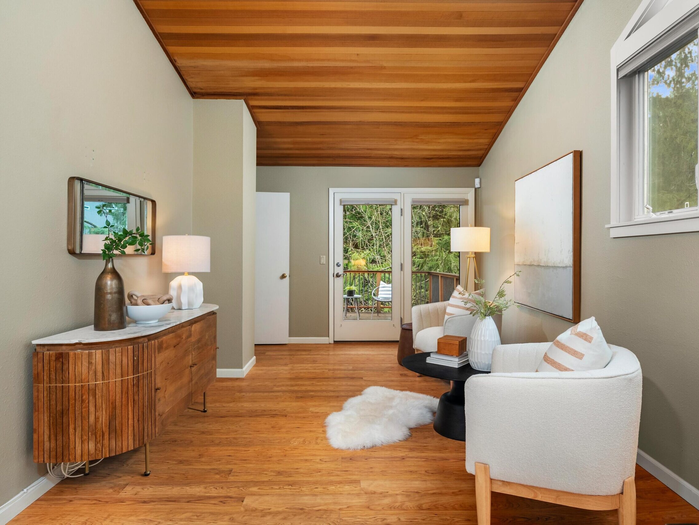 A cozy sitting area with two white armchairs, a round black table, and a white rug on wood flooring. A wooden sideboard with decor sits against the wall, with a mirror above and large windows letting in natural light.