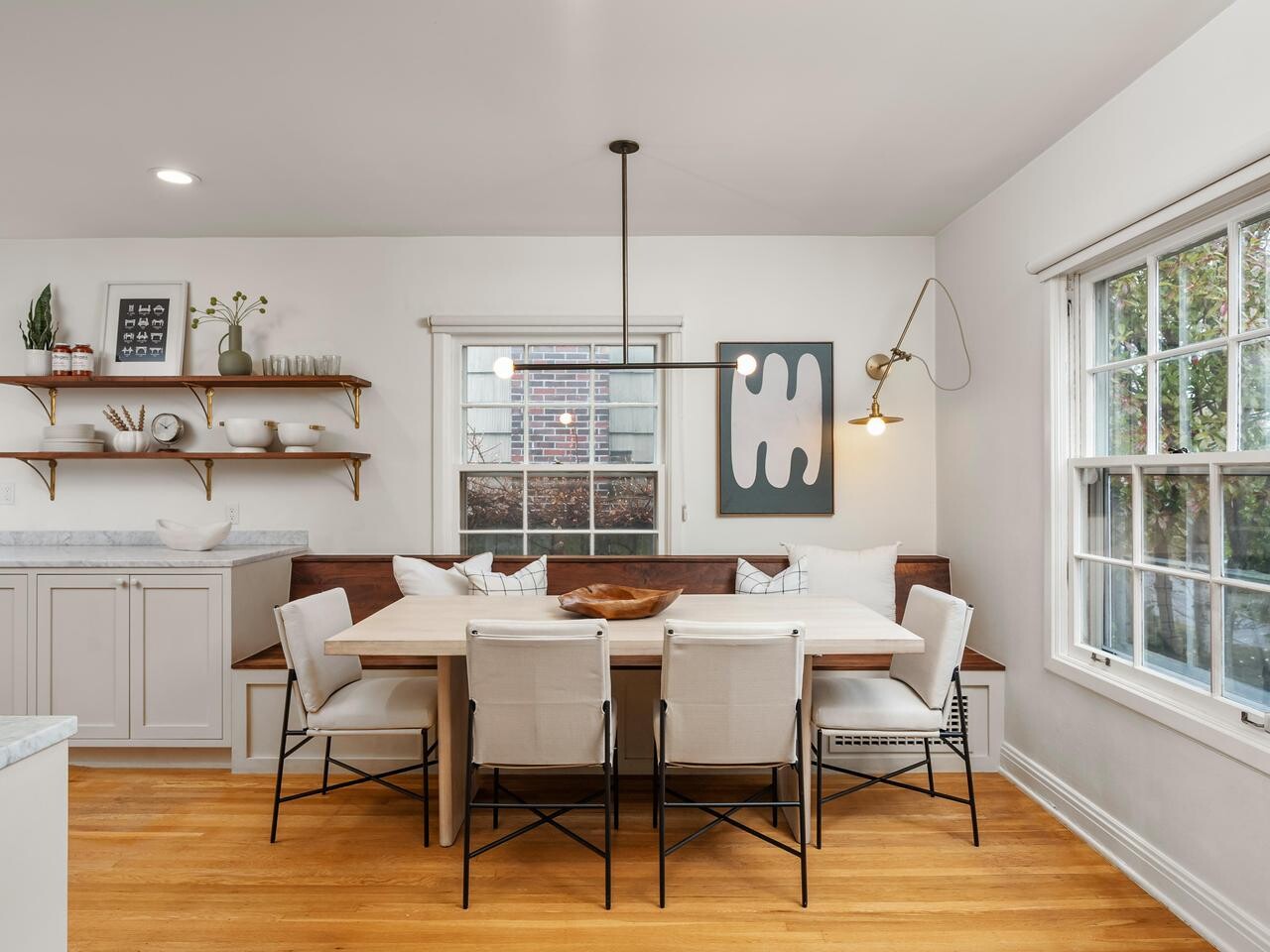 A bright, modern dining area with a wooden table, six white chairs, a built-in bench, minimalist decor, open shelves with dishes, and large windows letting in natural light.