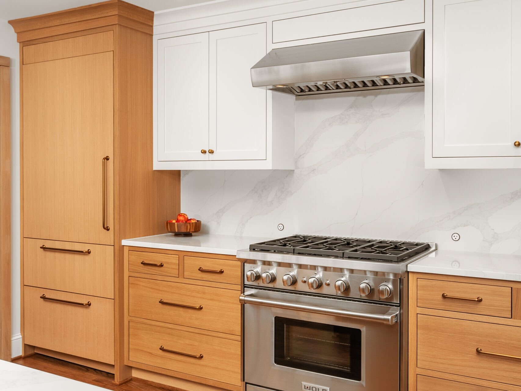 Modern kitchen with light wood cabinets, white marble countertops and backsplash, stainless steel gas stove with hood, and integrated wood-paneled refrigerator. Hardwood floors and recessed lighting enhance the clean, bright space.