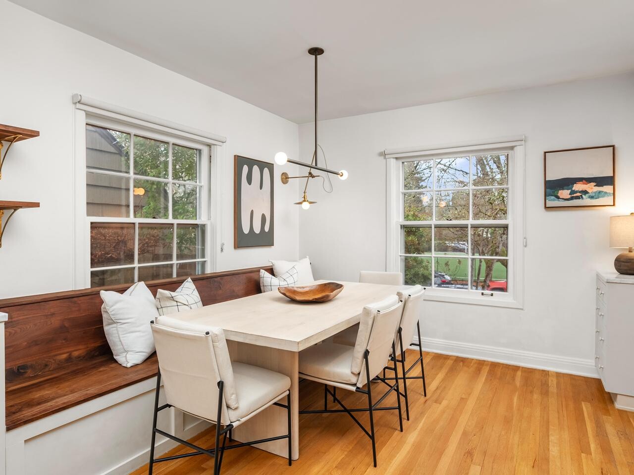 A cozy dining nook with a wooden bench, white chairs, and a rectangular table sits by two large windows. Minimalist decor, modern light fixture, art on the walls, and hardwood floors complete the inviting space.