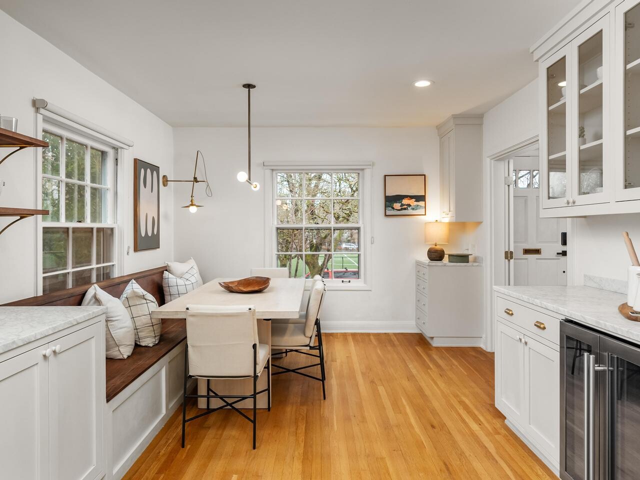 Bright, modern kitchen with wood floors, white cabinets, marble countertops, built-in bench seating around a dining table, minimalist decor, large windows, and pendant lighting.