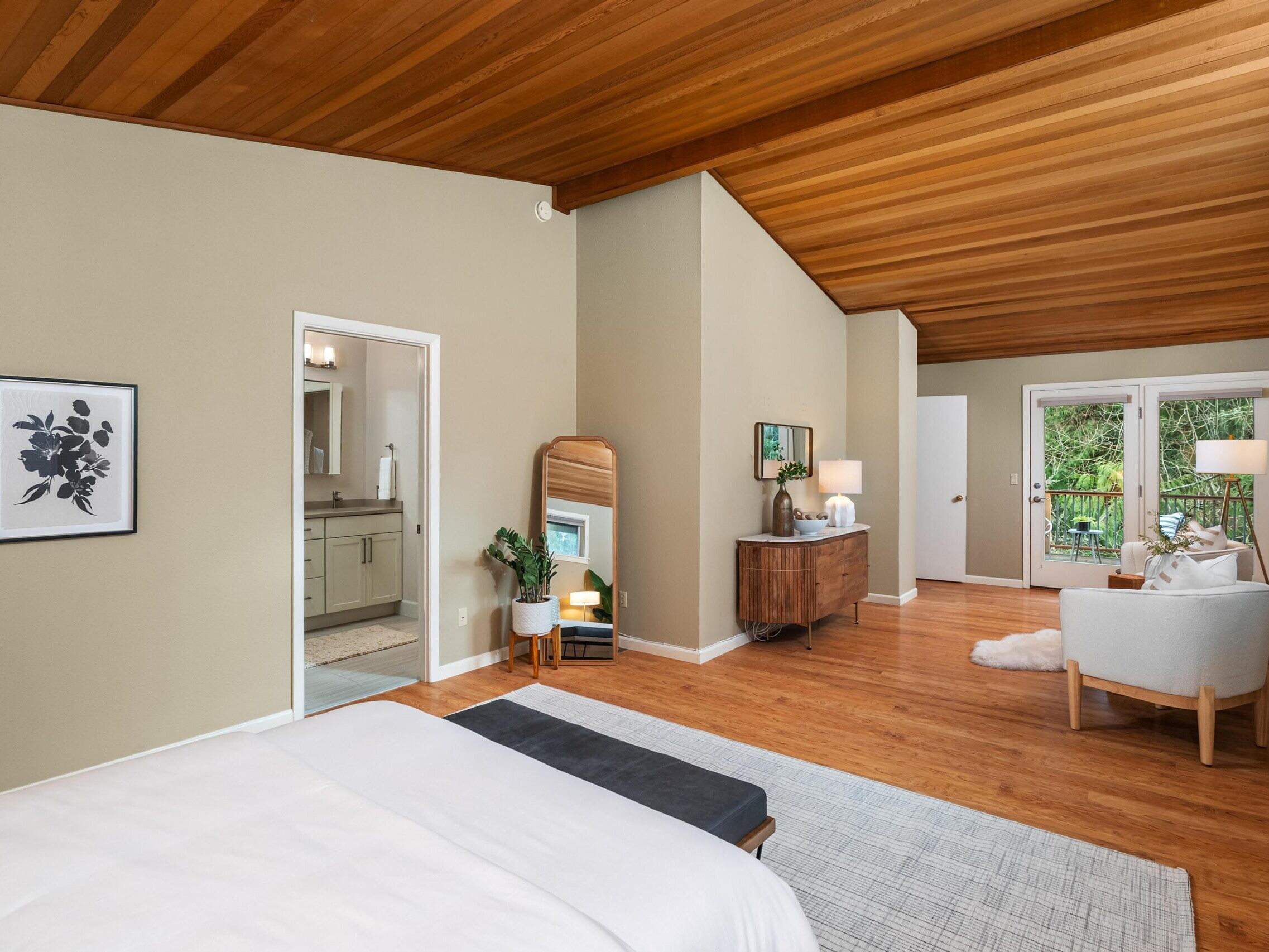 Spacious bedroom with wood-paneled vaulted ceiling, beige walls, and wood floors. Features a bed, armchair, dresser, and floor mirror. Sliding doors lead to a balcony with greenery outside. Bathroom visible through open door.