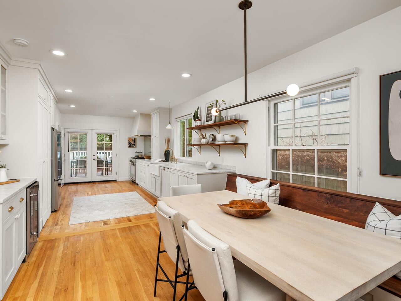 Bright, modern kitchen with white cabinets, wood floors, and a long dining table with cushioned chairs and a wooden bench. Large windows let in natural light, and open shelves hold decor and dishes.