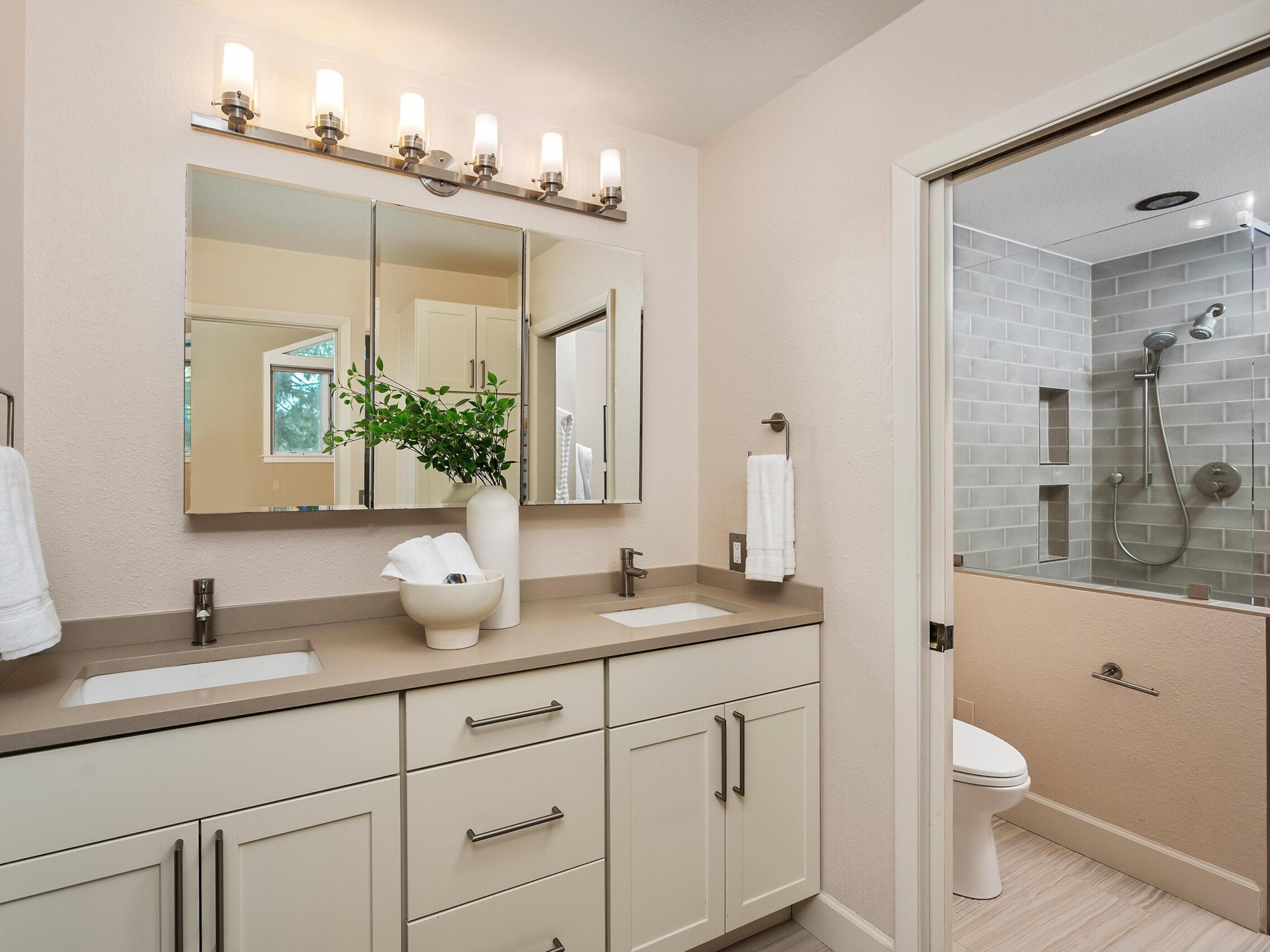 Modern bathroom with a double-sink vanity, large mirrors, wall sconces, white cabinets, and a vase with greenery. In the background, a shower with gray tiles and a glass door, plus a toilet are visible.