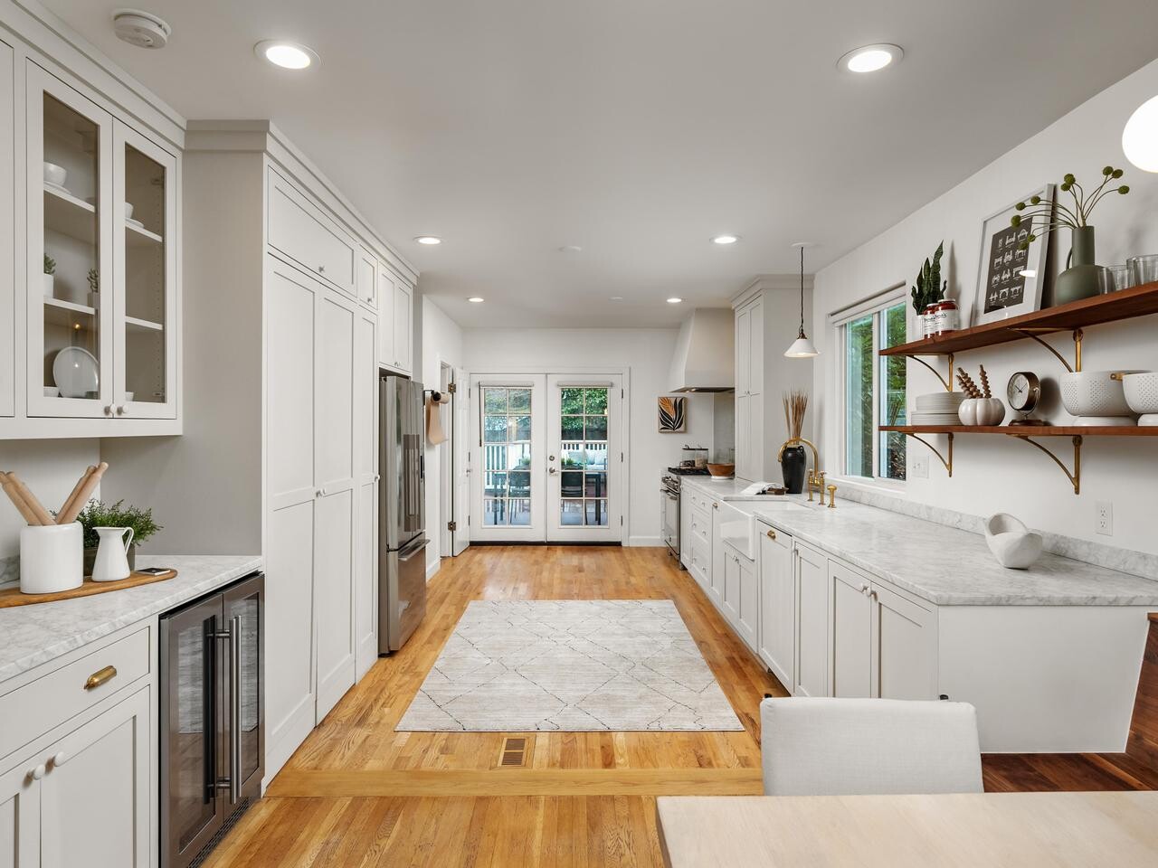 Modern kitchen with white cabinets, marble countertops, stainless steel appliances, open wooden shelves with dishes and plants, hardwood floors, and glass doors leading to a patio. A dining area is partially visible in the foreground.
