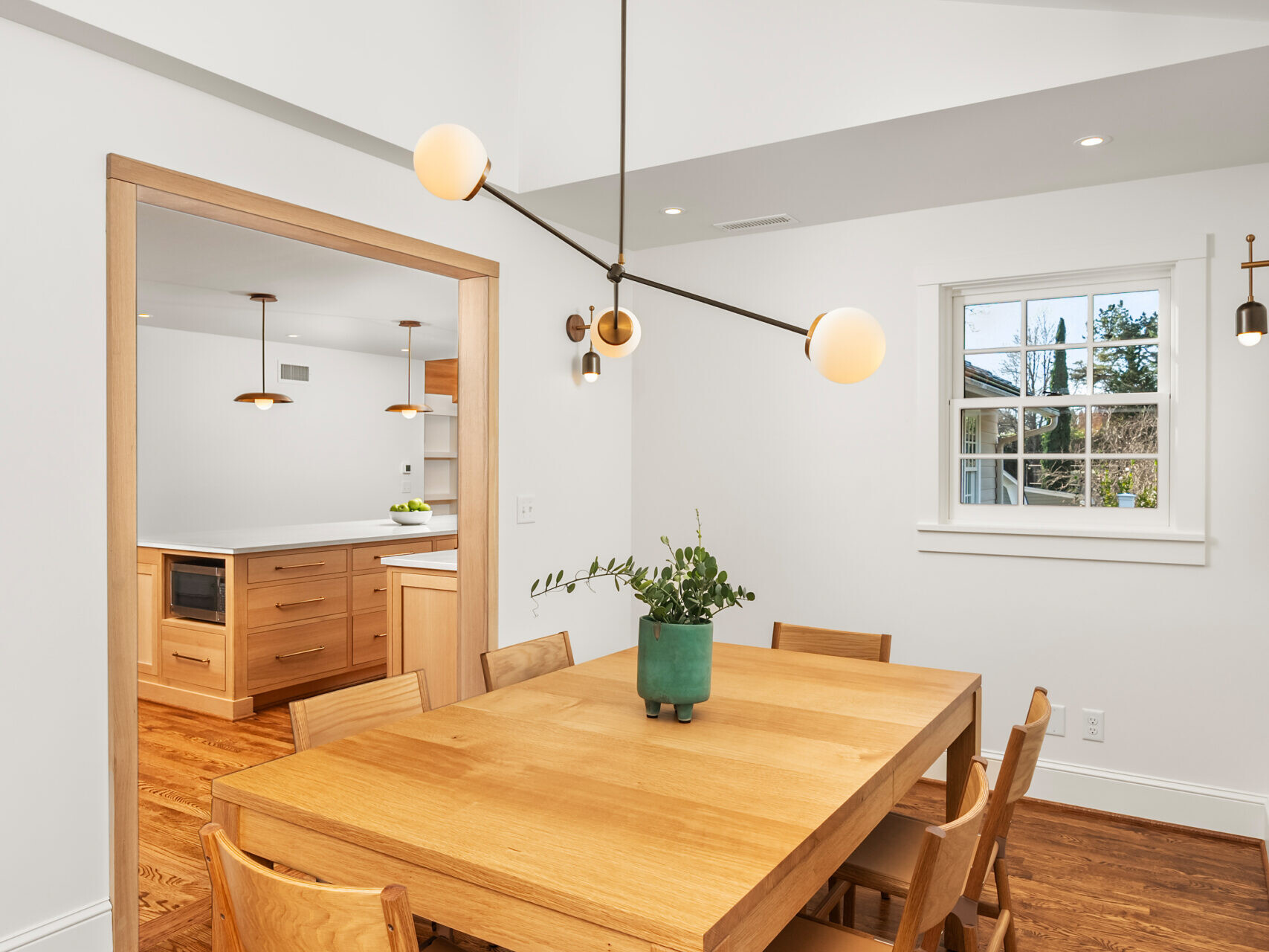 A modern dining room with a wooden table, six matching chairs, a potted plant centerpiece, hardwood floors, white walls, and a contemporary ceiling light. The adjacent kitchen is visible through an open doorway.