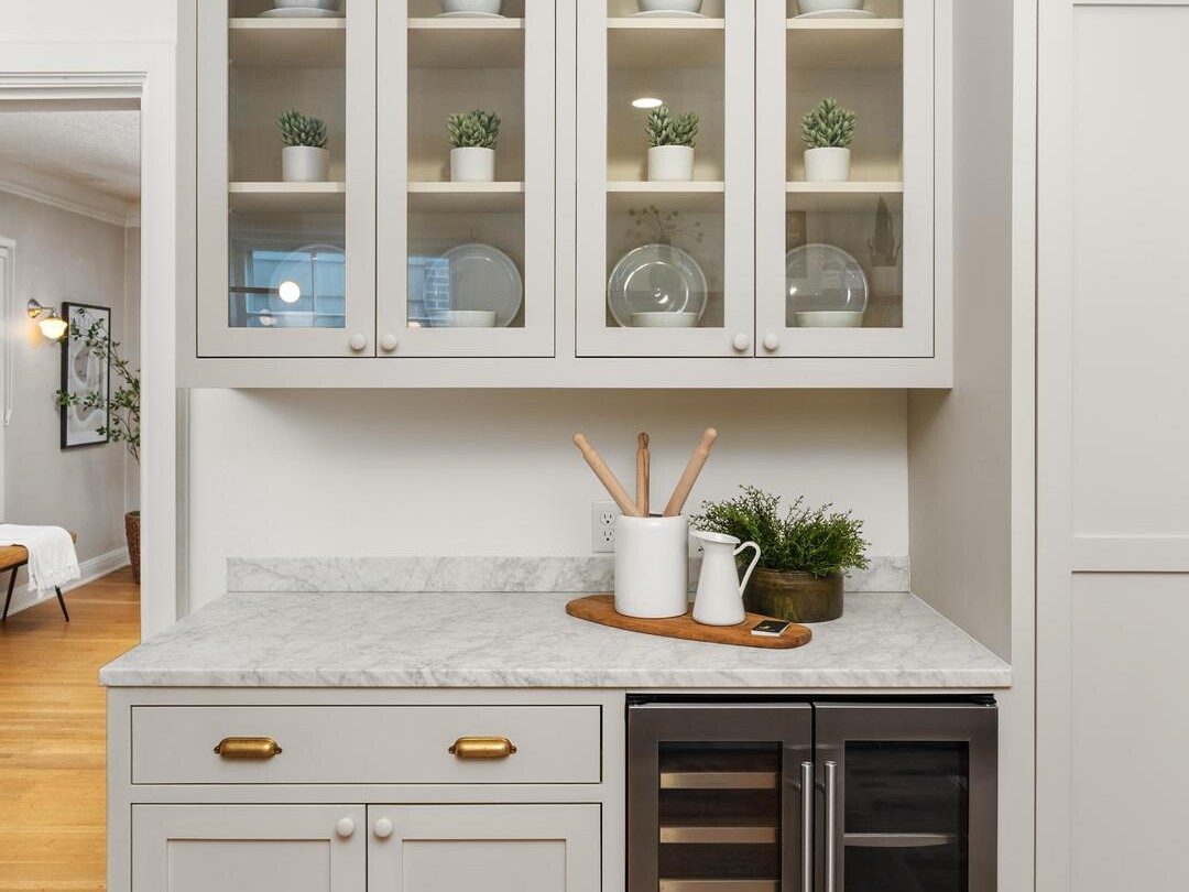 A modern kitchen corner with glass-front cabinets holding plates and plants, marble countertop with utensils and decor, and a built-in dual wine fridge below. The floor is wooden and the overall style is clean and minimal.