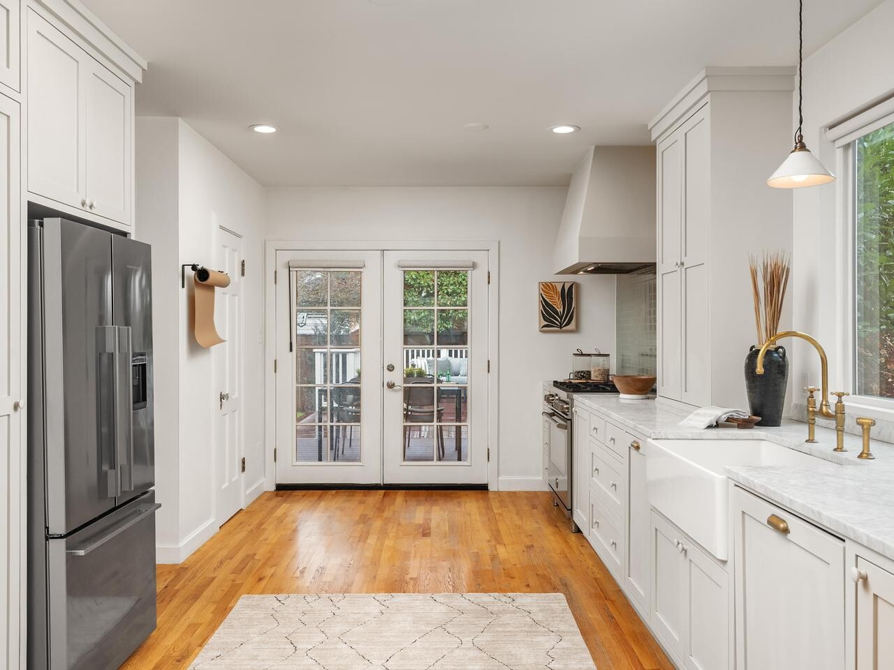 Bright, modern kitchen with white cabinets, marble counters, stainless steel appliances, light wood flooring, a rug, and double glass doors leading to an outdoor deck with patio furniture.