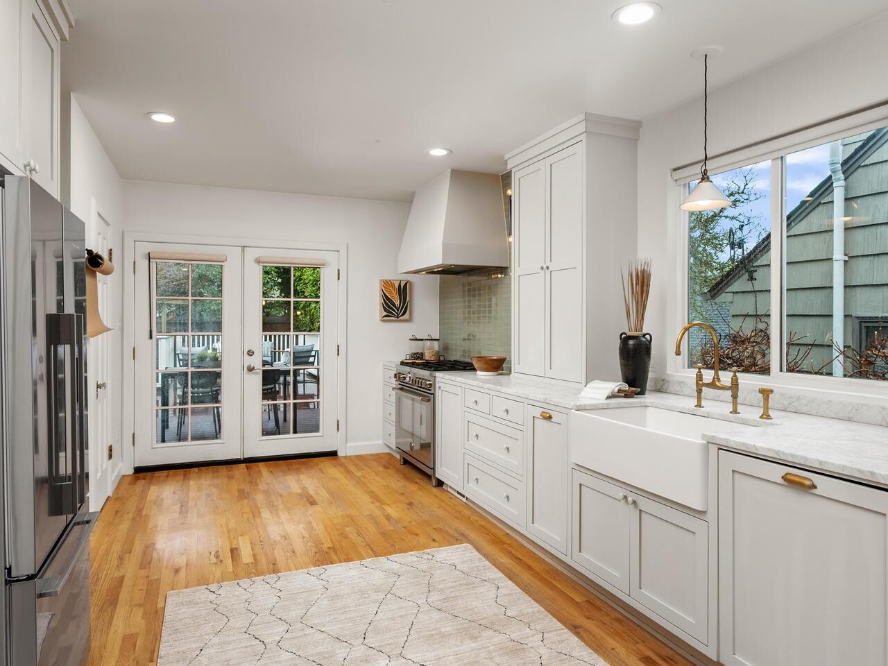 Bright, modern kitchen with white cabinets, marble countertops, stainless steel appliances, a farmhouse sink, and hardwood floors. Glass doors lead to a patio, and a window lets in natural light above the sink.