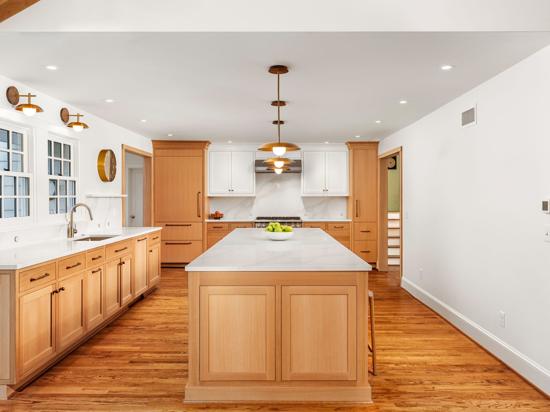 Spacious modern kitchen with wood cabinets, a large island with white countertop, hardwood floors, stainless steel appliances, and pendant lighting. White walls and ceiling create a bright, open atmosphere.