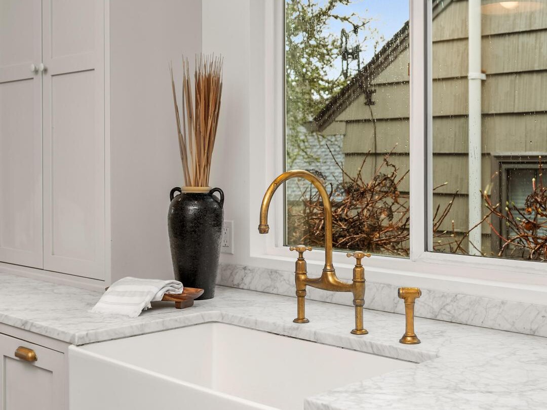 A modern kitchen with a white farmhouse sink, brass faucet, marble countertops, light grey cabinets, a decorative black vase with dried reeds, and a window showing the exterior of a house and blue sky.