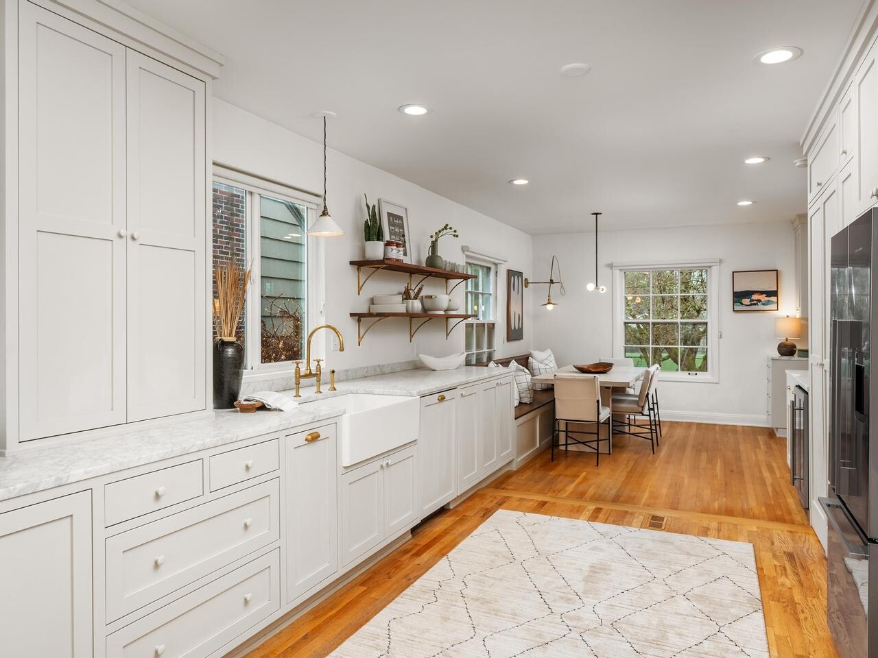Bright, modern kitchen with white cabinets, marble countertops, a farmhouse sink, open wooden shelves, wood flooring, dining table with chairs, and large windows letting in natural light.