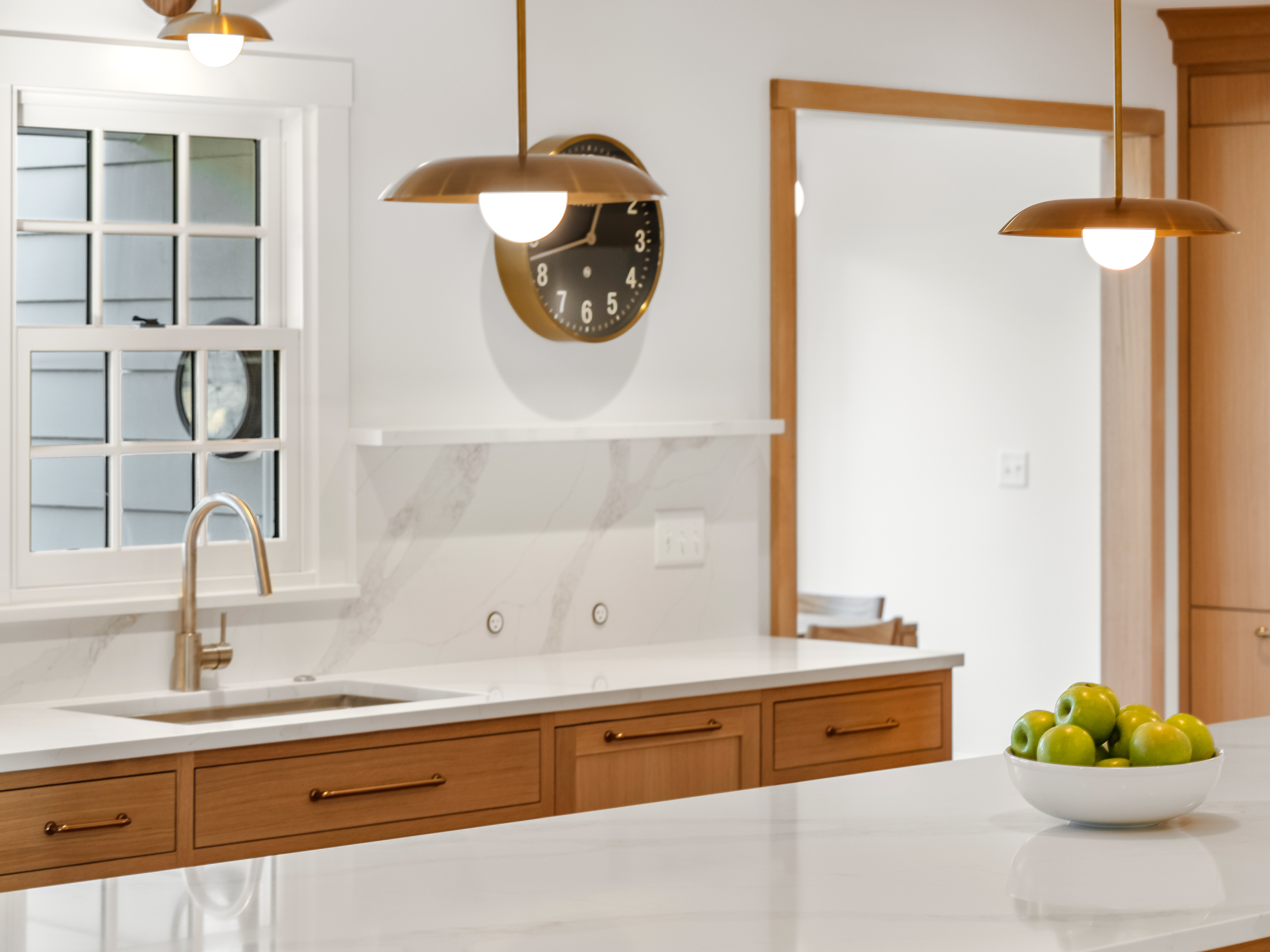 A modern kitchen with wood cabinets, a white marble countertop, a sink under a window, pendant lights, a wall clock, and a bowl of green apples on the island. The space is bright and minimalistic.