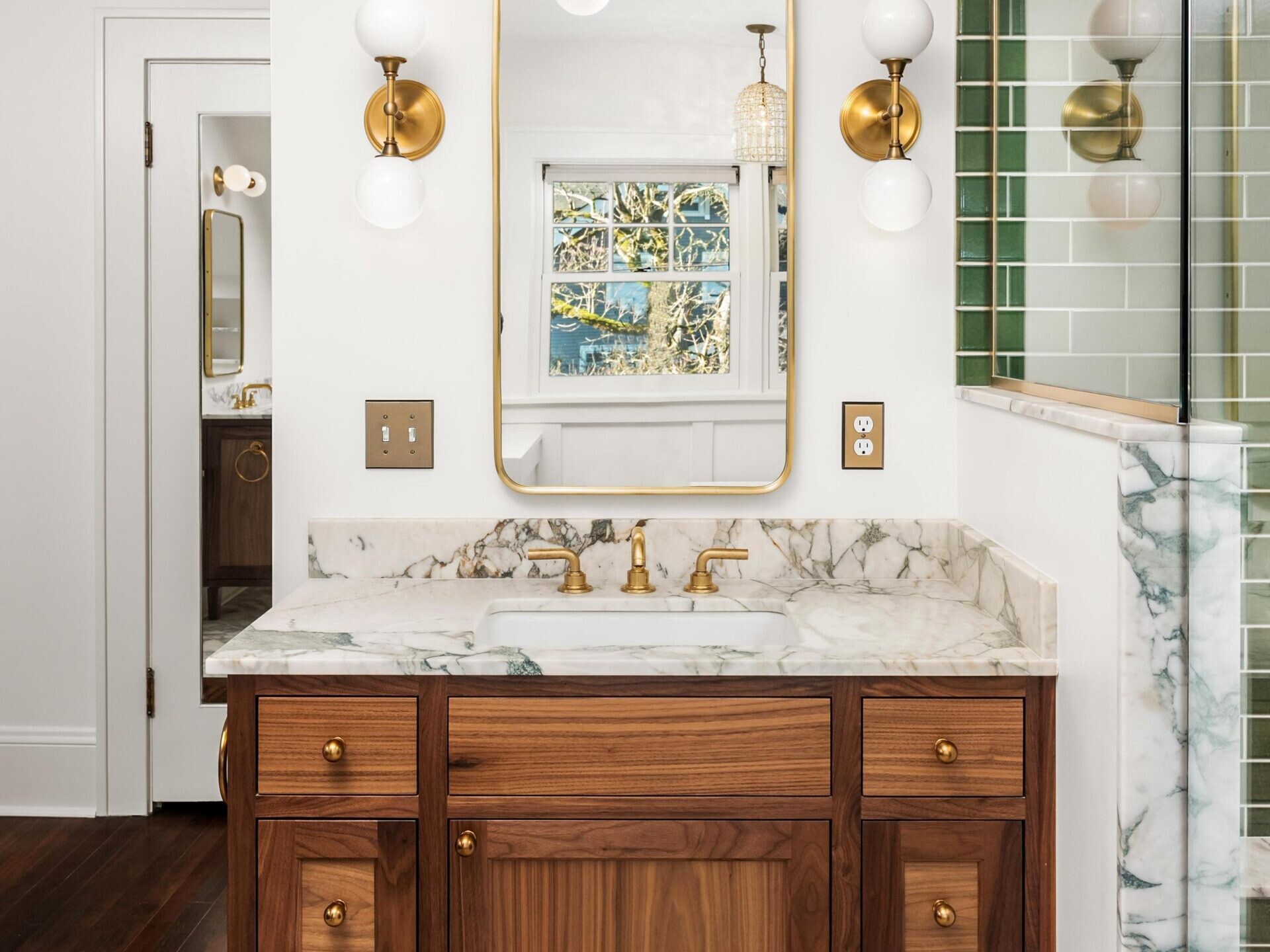 A bathroom vanity with a wood cabinet, marble countertop, gold fixtures, a rectangular mirror with gold trim, and two wall sconces. A shower with green tile and glass walls is partially visible on the right.
