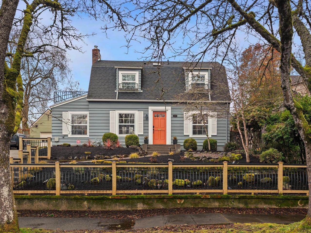 A two-story blue-gray house with white trim and an orange front door, set behind a wooden fence with a landscaped front yard featuring shrubs, rocks, and trees. The sky is clear and blue.