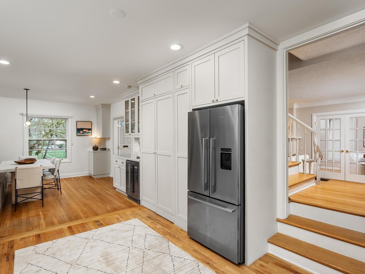 Bright, modern kitchen with white cabinets, stainless steel refrigerator, light wood floors, and a dining area by a window. A staircase with white railing leads up to a room with French doors.