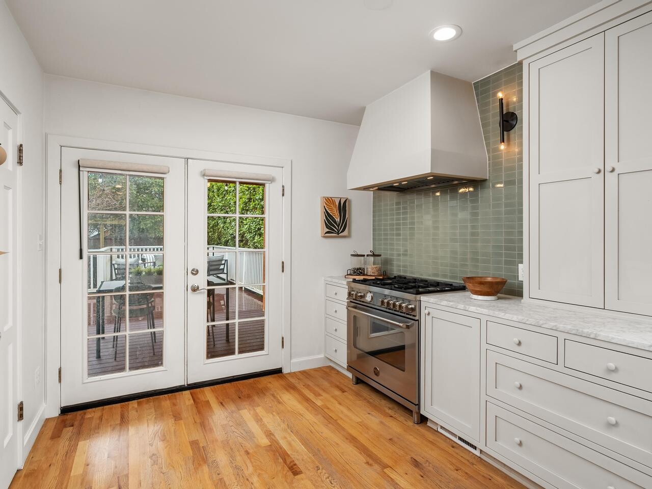 Bright kitchen with white cabinets, marble countertops, and light wood floors. Glass French doors open to a deck outside. Stainless steel stove with a vent hood sits against a pale green tile backsplash.