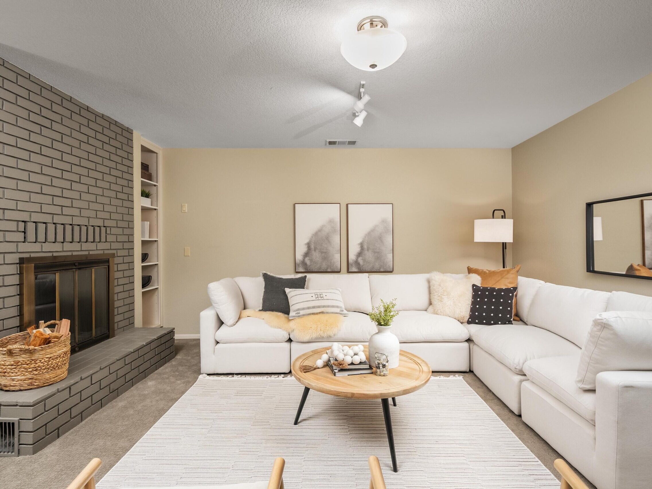 A modern living room with a white sectional sofa, neutral cushions, a round wooden coffee table, and a gray brick fireplace. Art pieces and a basket of firewood add decorative touches to the cozy, well-lit space.